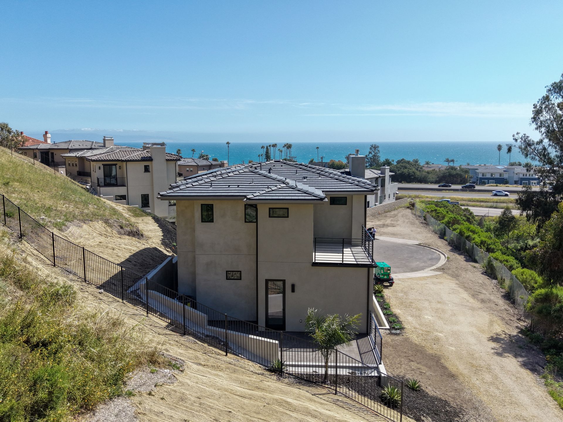 Residential houses on a hillside overlooking the ocean, with a dirt road and clear blue sky.