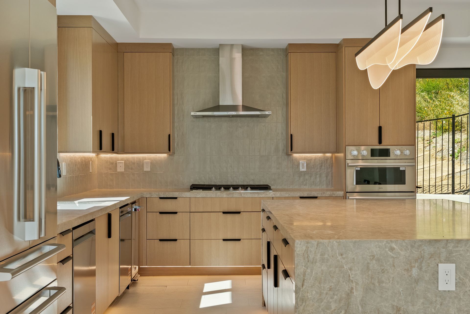 Modern kitchen with light wood cabinets, marble island, stainless appliances, and large windows