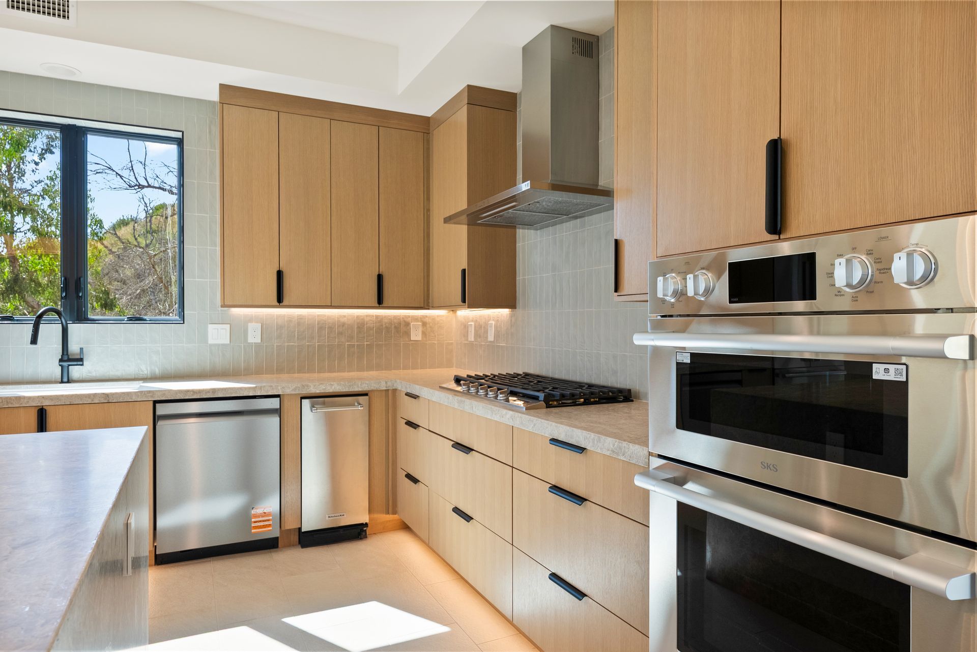 Modern kitchen with light wood cabinets, stainless steel appliances, and a window over the sink.