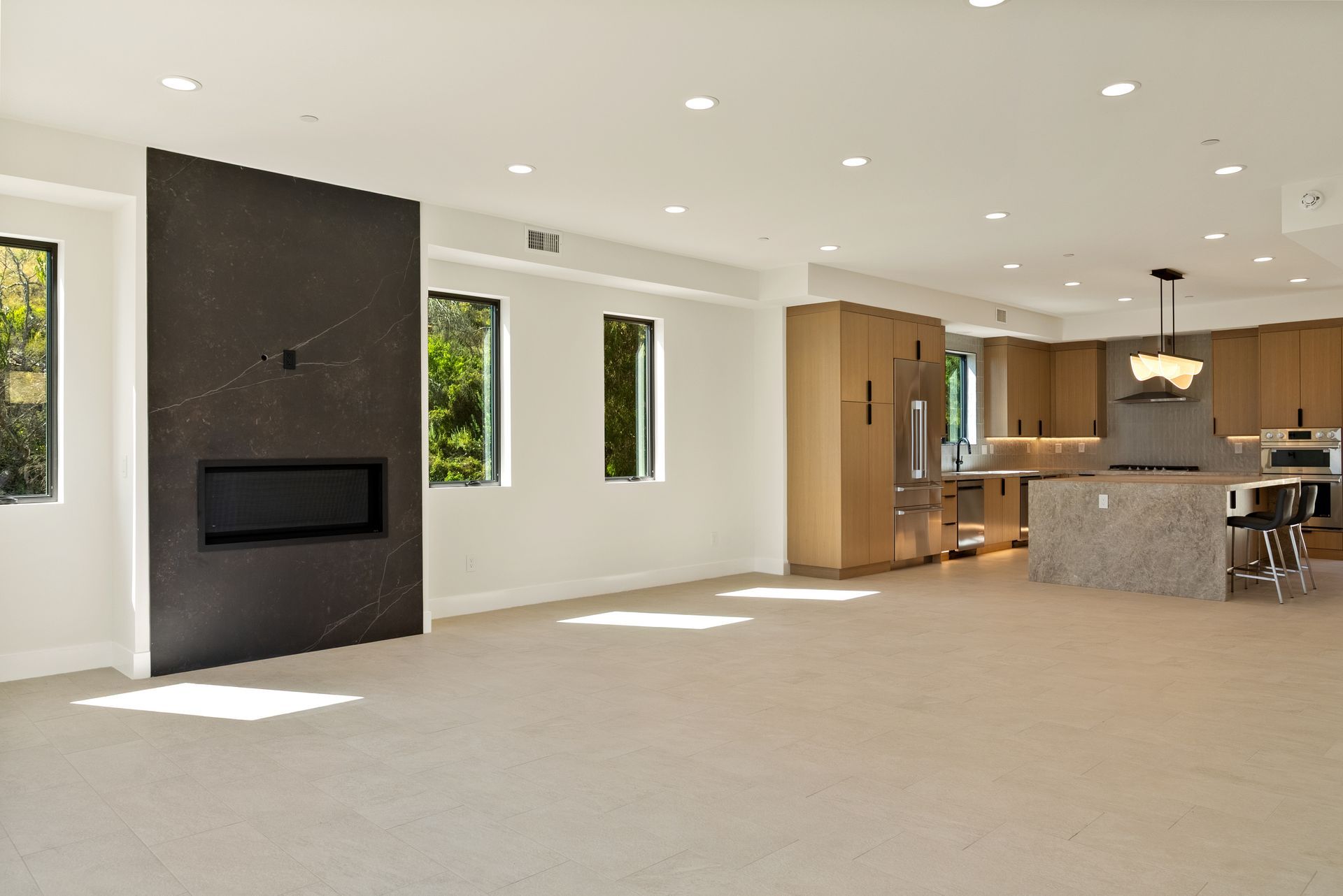 Spacious empty living room with beige carpet, dark fireplace wall, large windows, and an open kitchen.