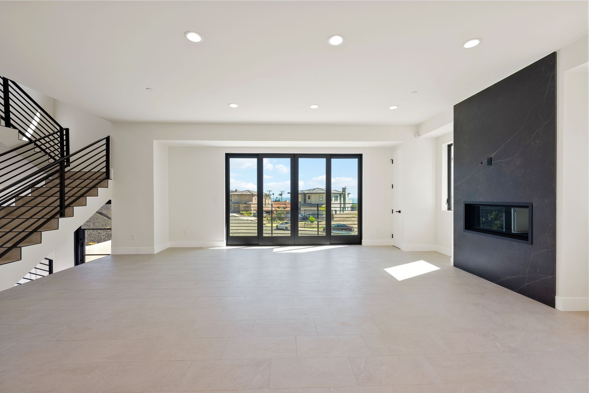 Bright open living room with white walls, carpet, black staircase, and fireplace, facing large glass doors.