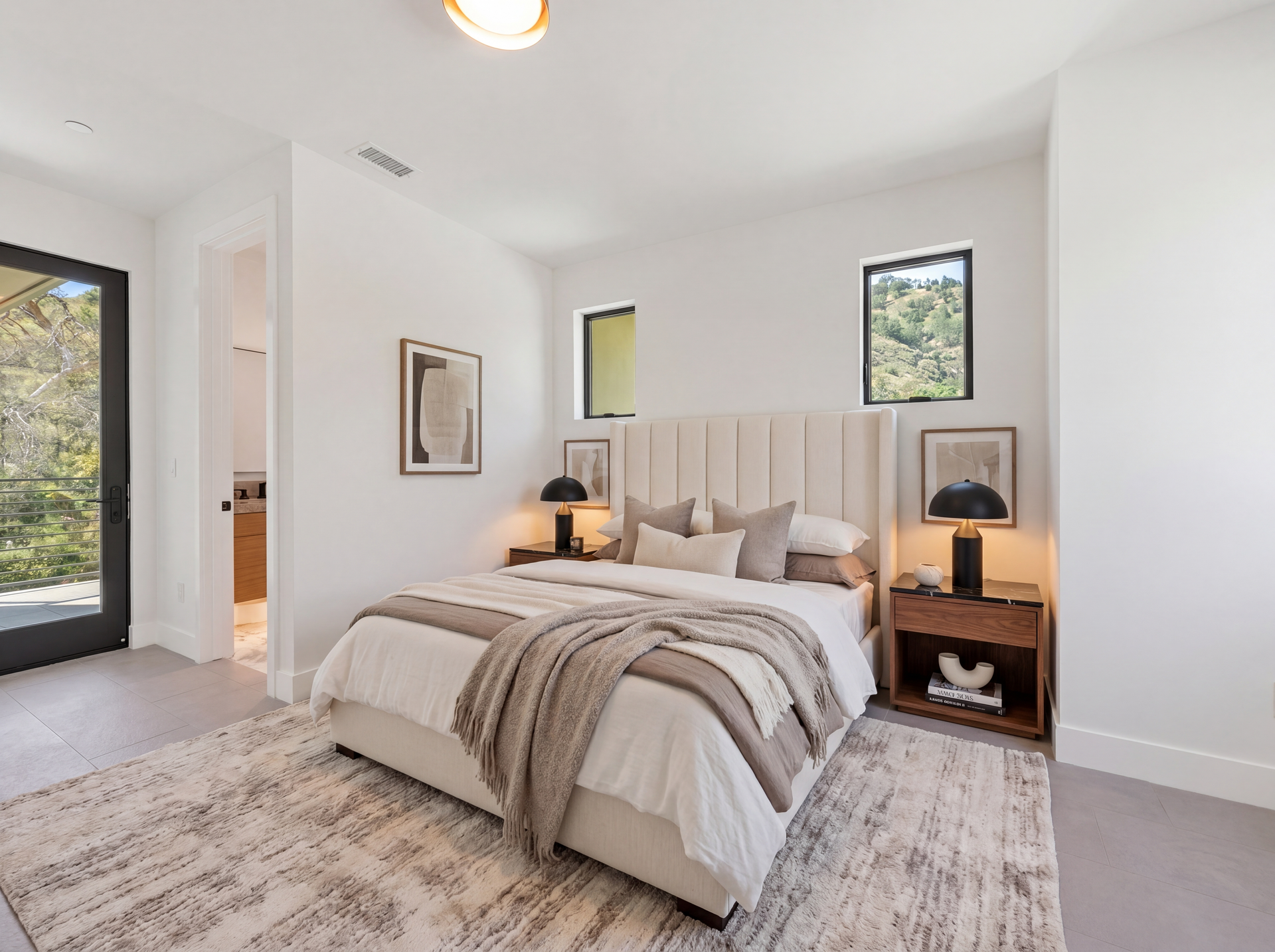 Bright bedroom with a neatly made bed, bedside lamps, and white walls with framed art and windows.