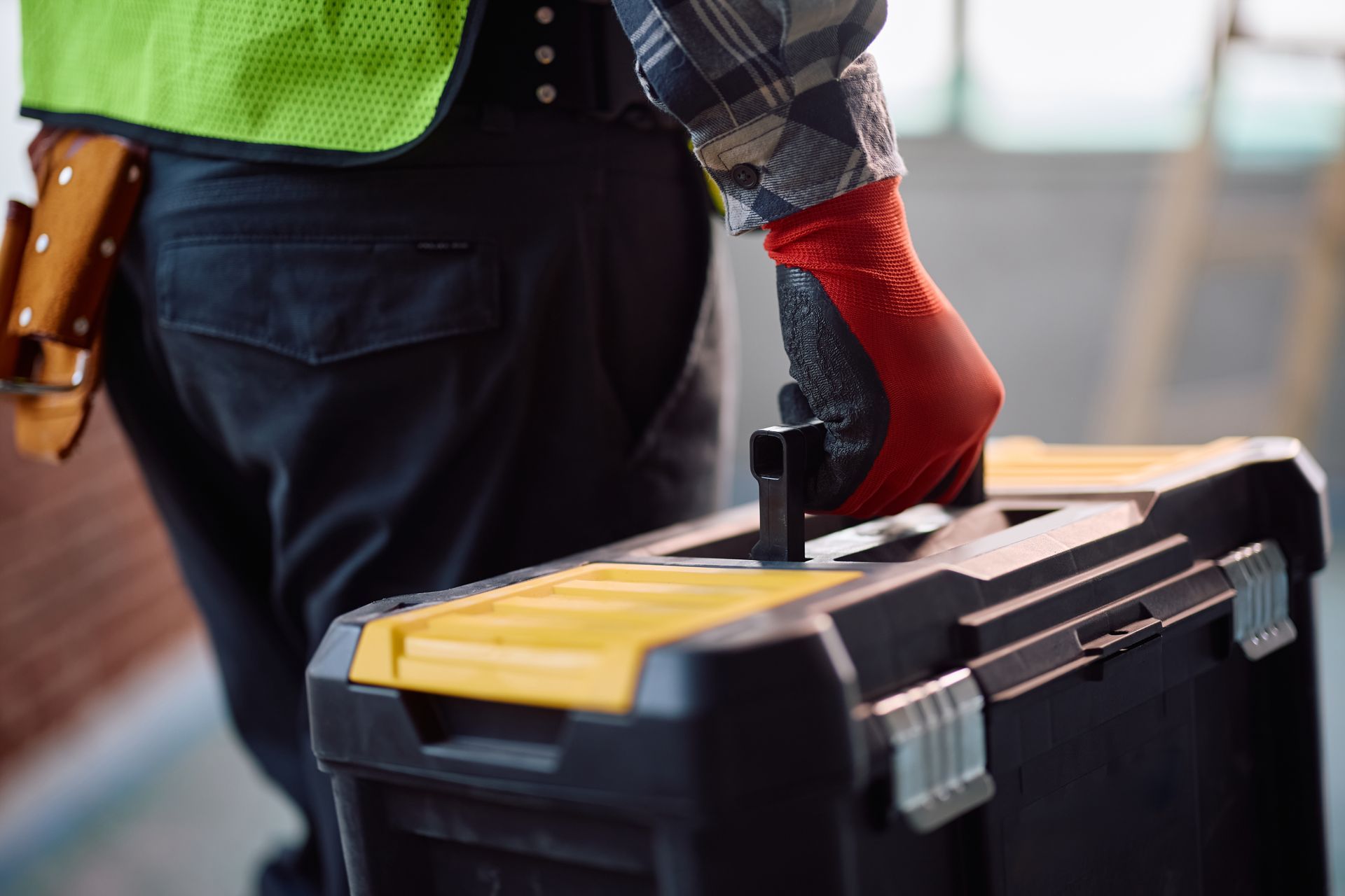 Construction worker carrying a black toolbox, wearing a green vest and red gloves outdoors