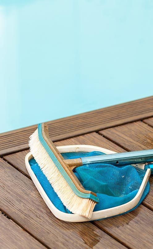 A brush and net are sitting on a wooden deck next to a swimming pool.