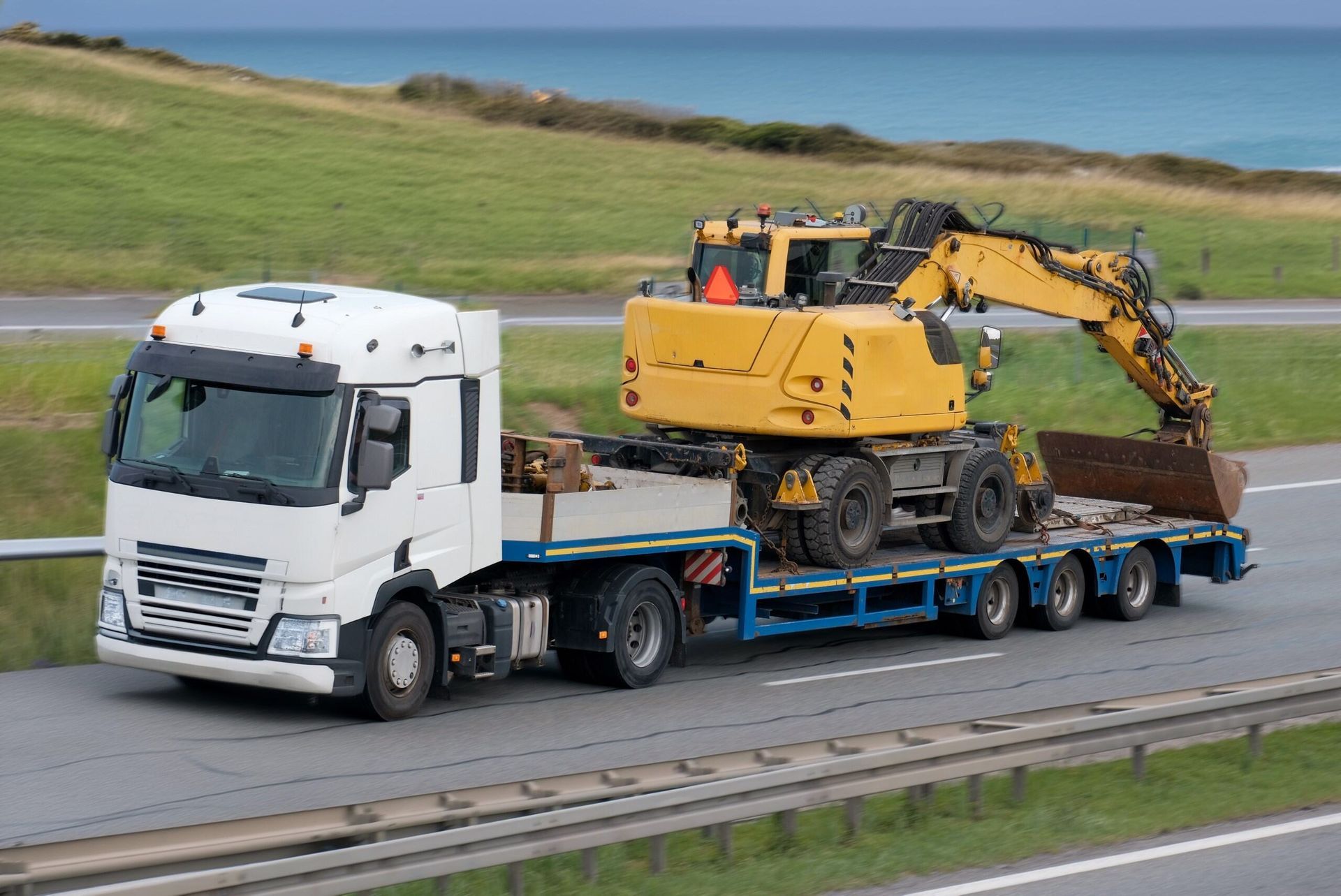 A White Truck With a Yellow Excavator on the Back is Driving Down a Highway — Hervey Bay Tilt Tray Hire In Maryborough, QLD