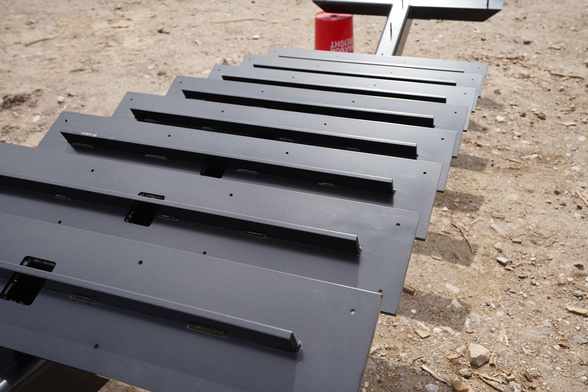 Black metal panels laid out outdoors on a dirt surface, viewed in perspective.