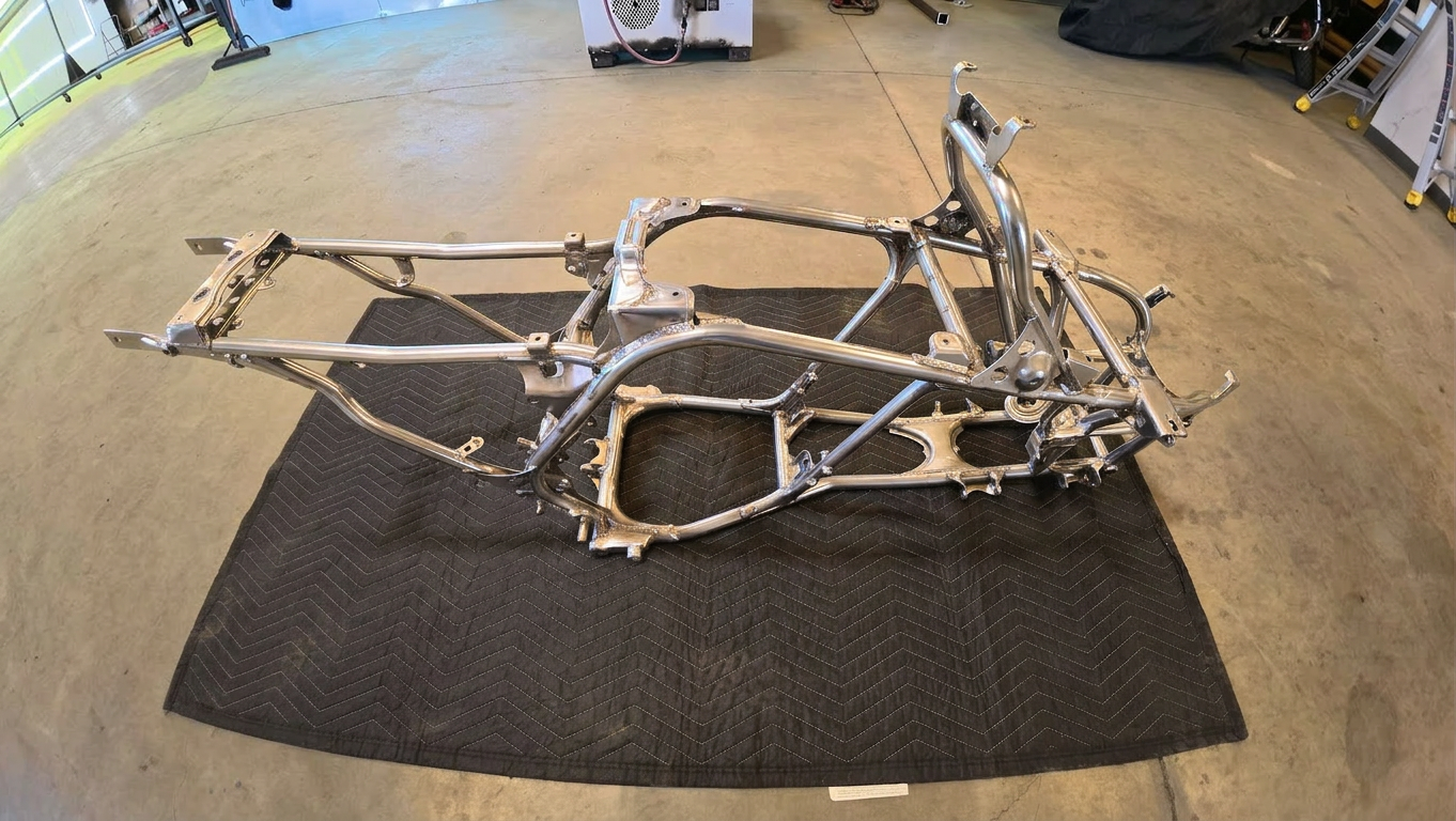 Bare metal motorcycle frame on a black mat in a workshop, viewed from above.