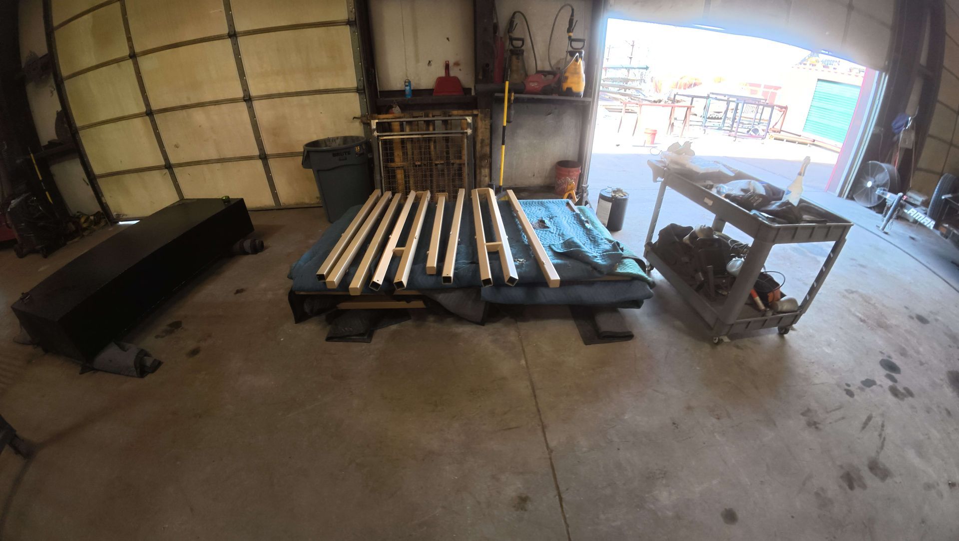 Workshop with stacked wooden pallets, a black bench, and a table saw near an open garage door.