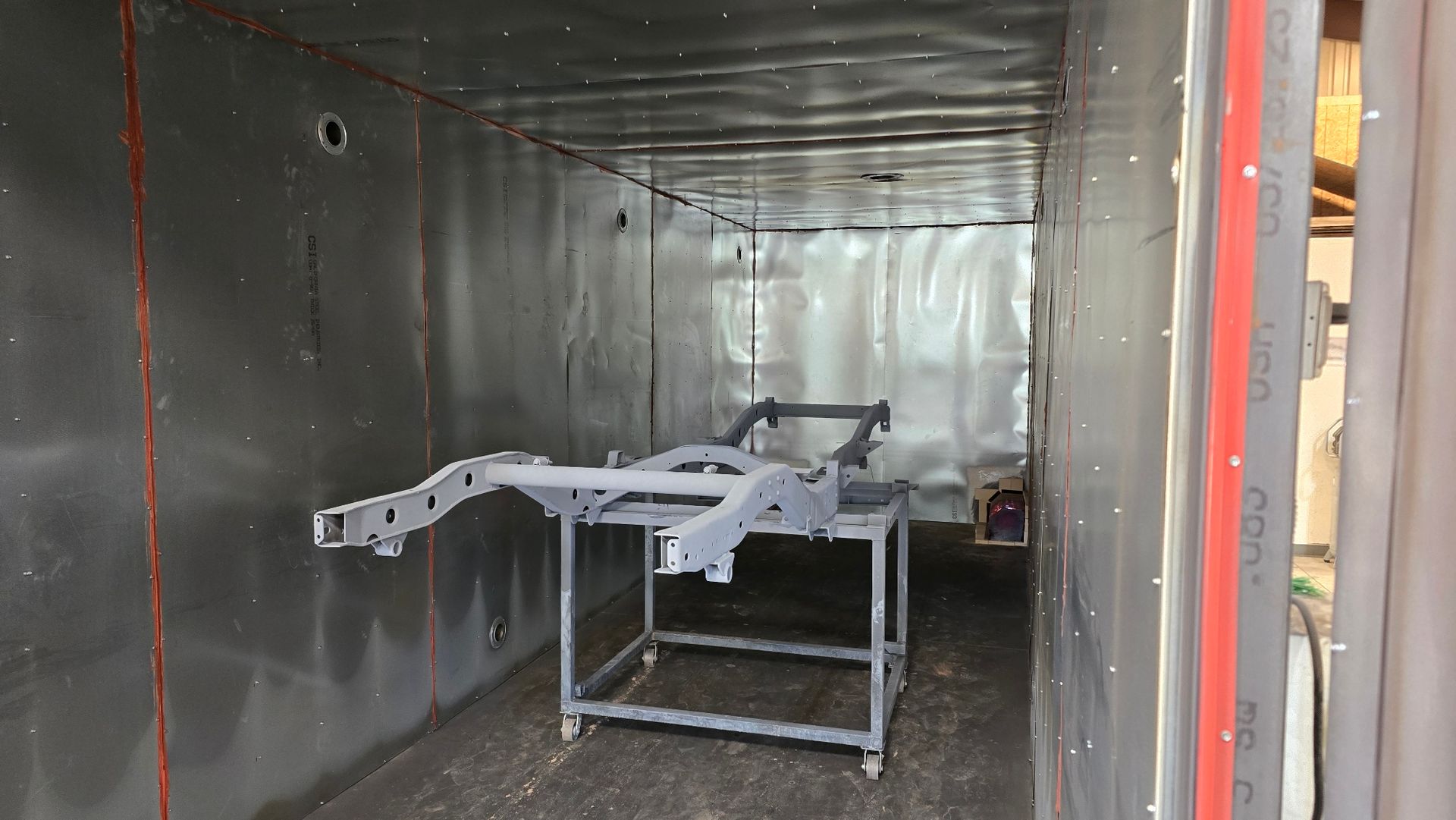 Metal machine frame in a silver-walled workshop or storage room, with a red door frame at right.