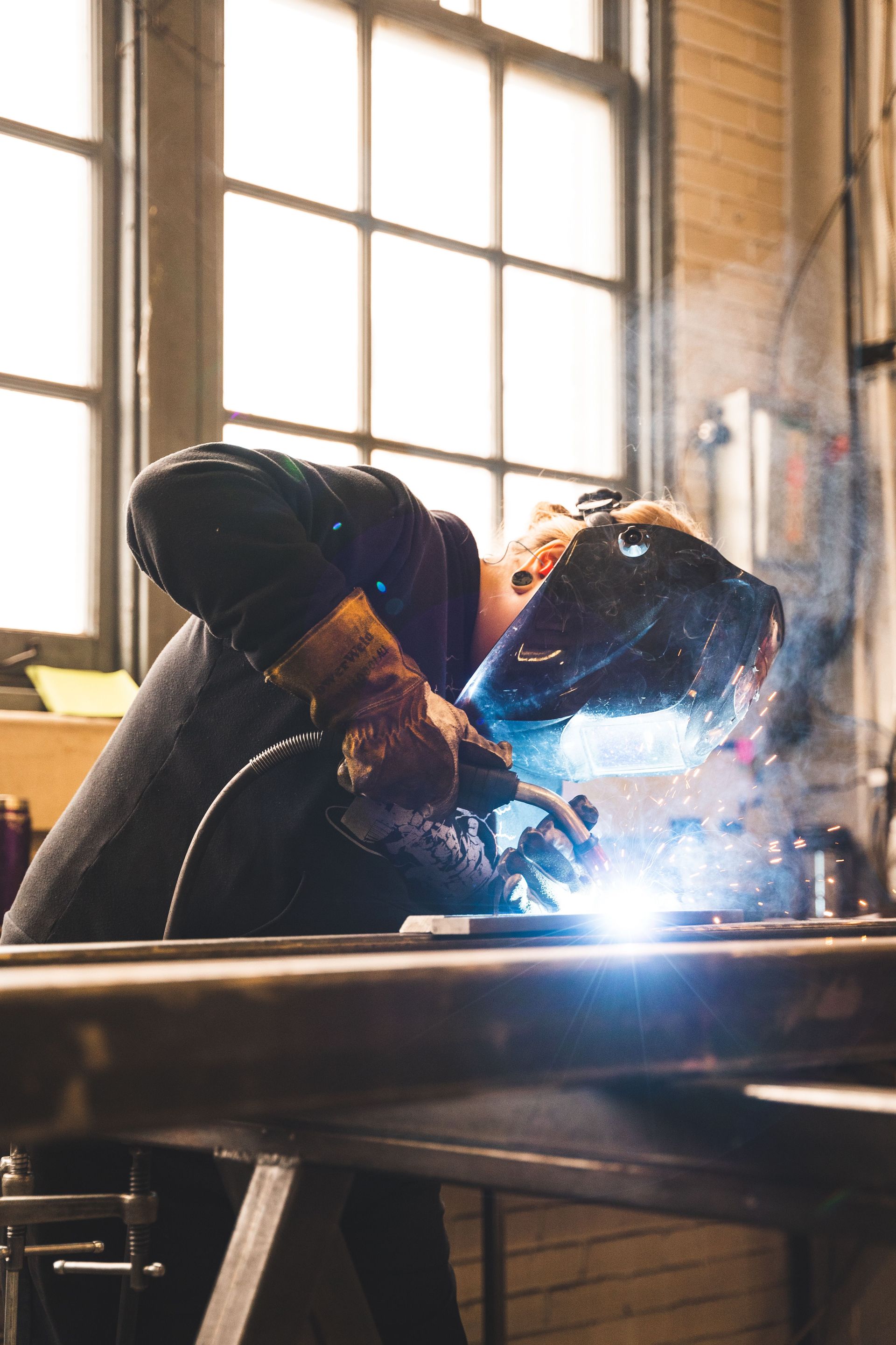 Welding worker in a shop, sparks flying beside a bright window