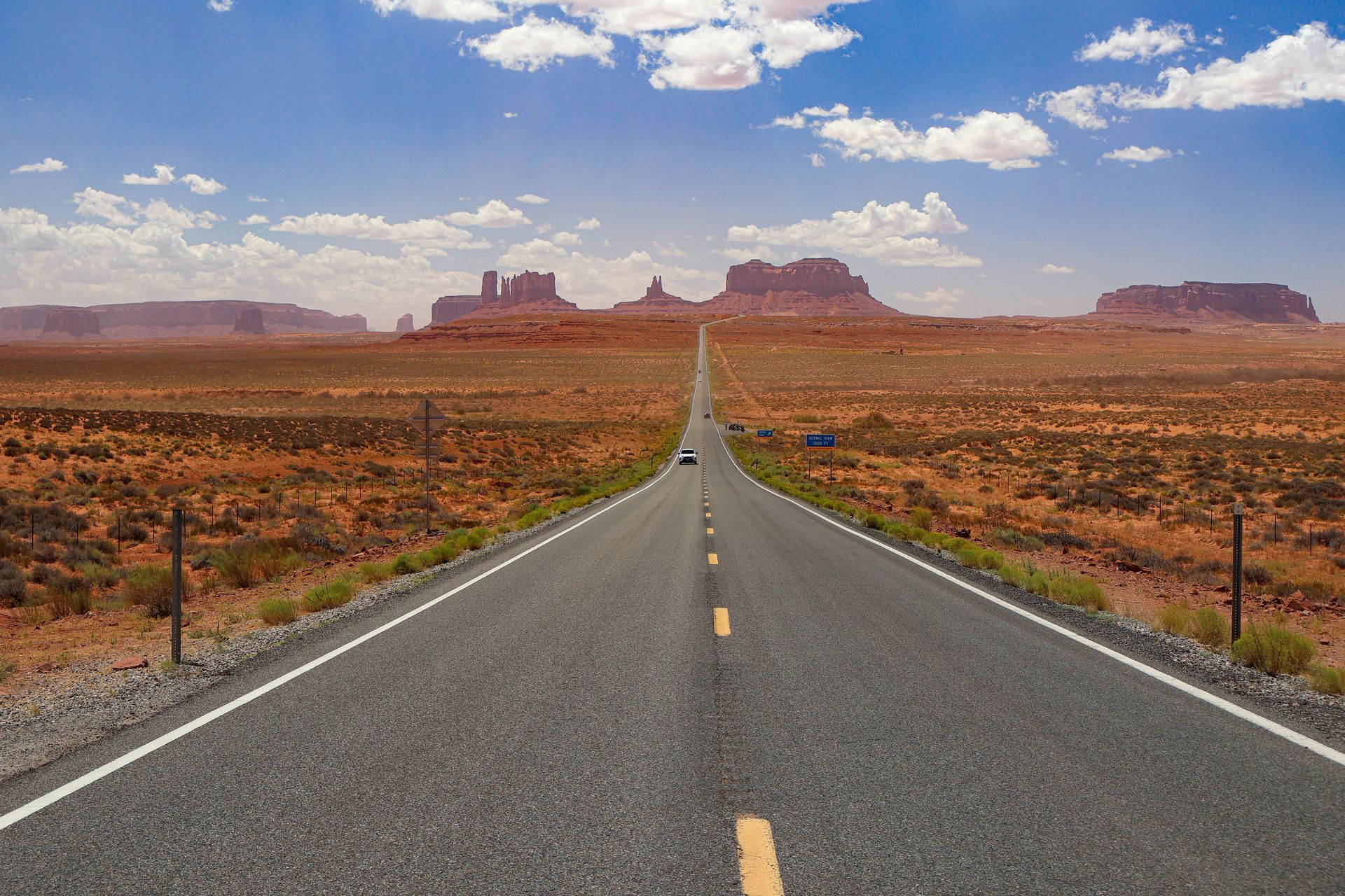 Straight desert highway leading toward distant red rock mesas under a blue, partly cloudy sky