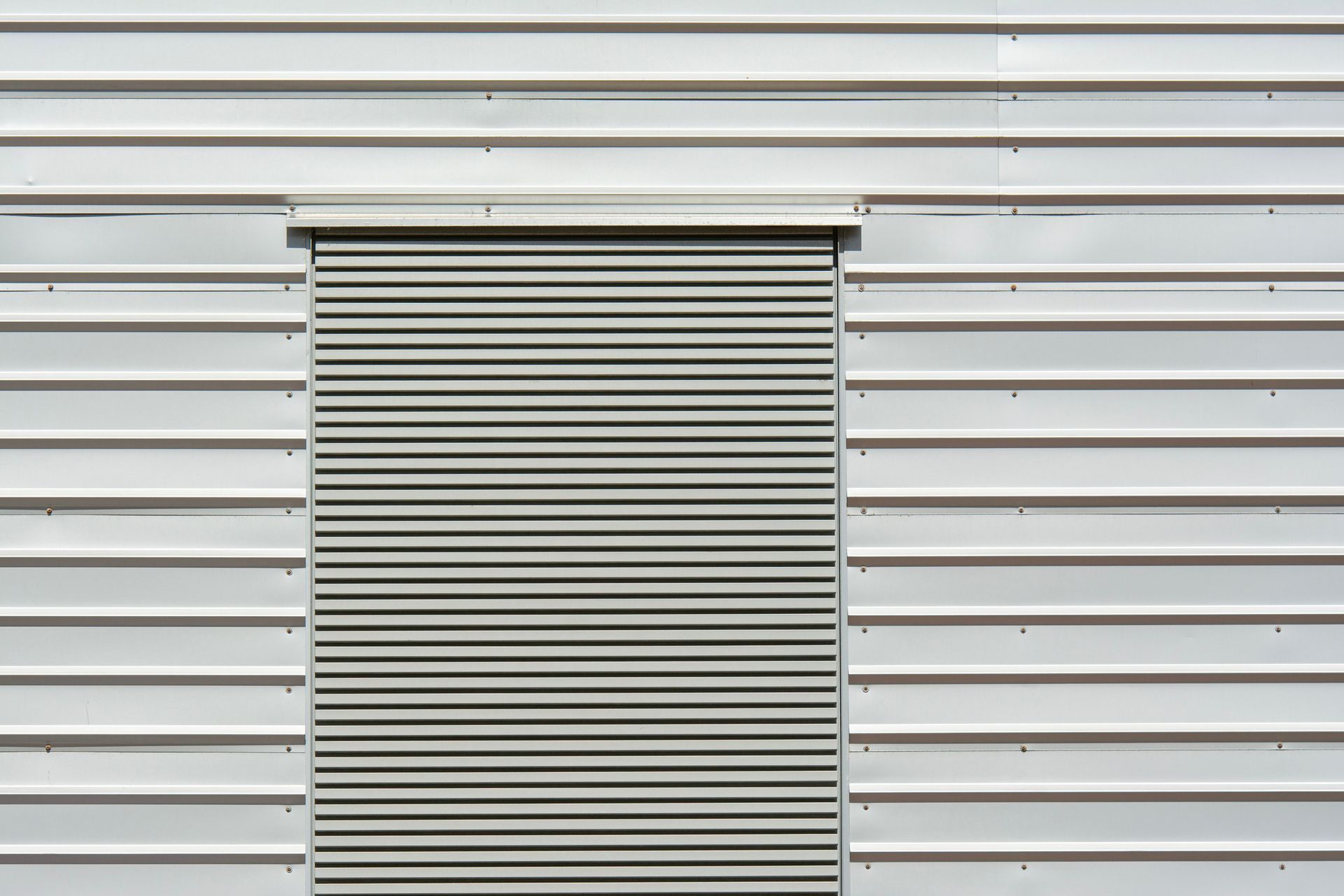 White metal building wall with a closed gray roll-up shutter door centered in the frame