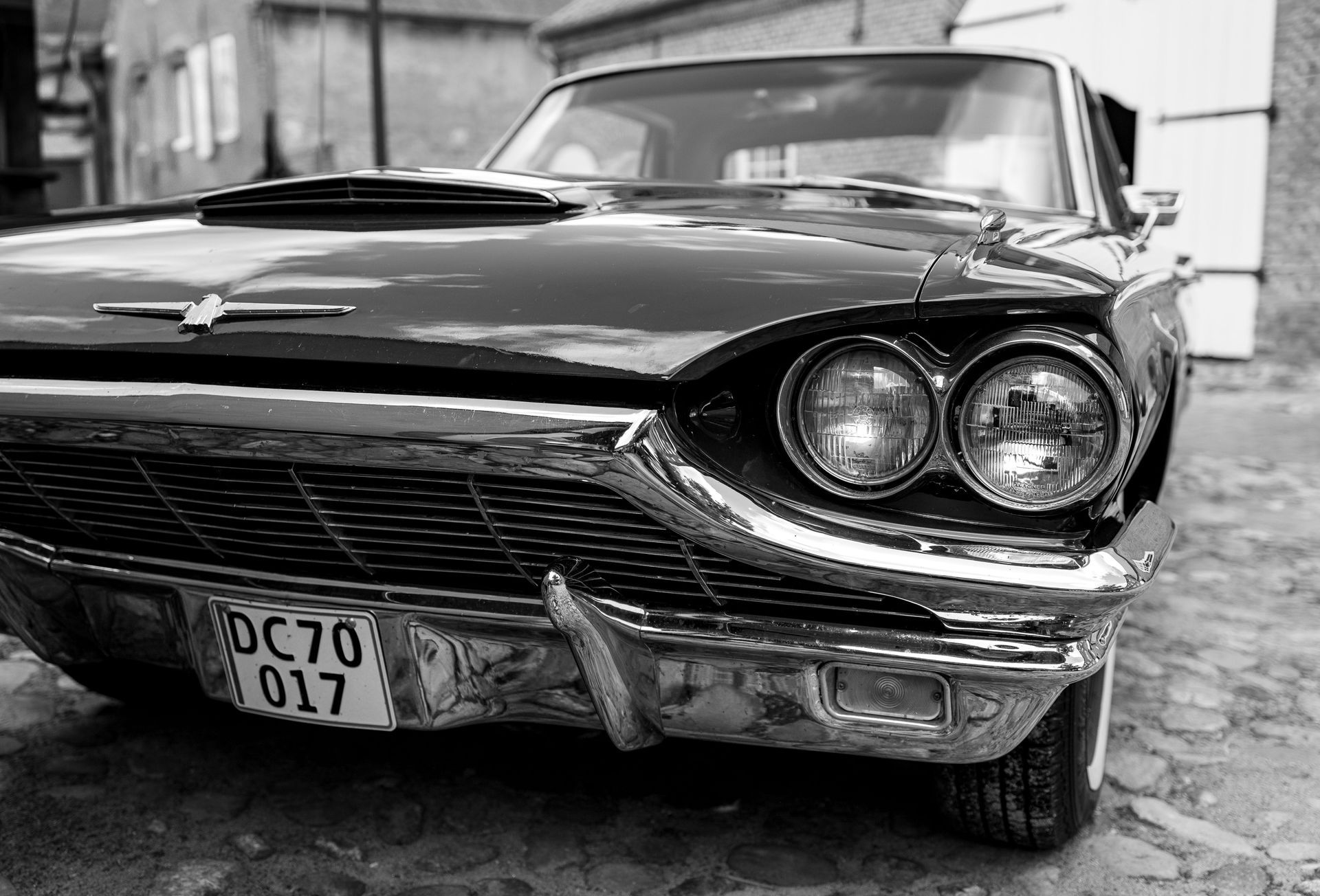 Black-and-white close-up of a vintage car’s front end with round headlights and a visible license plate.