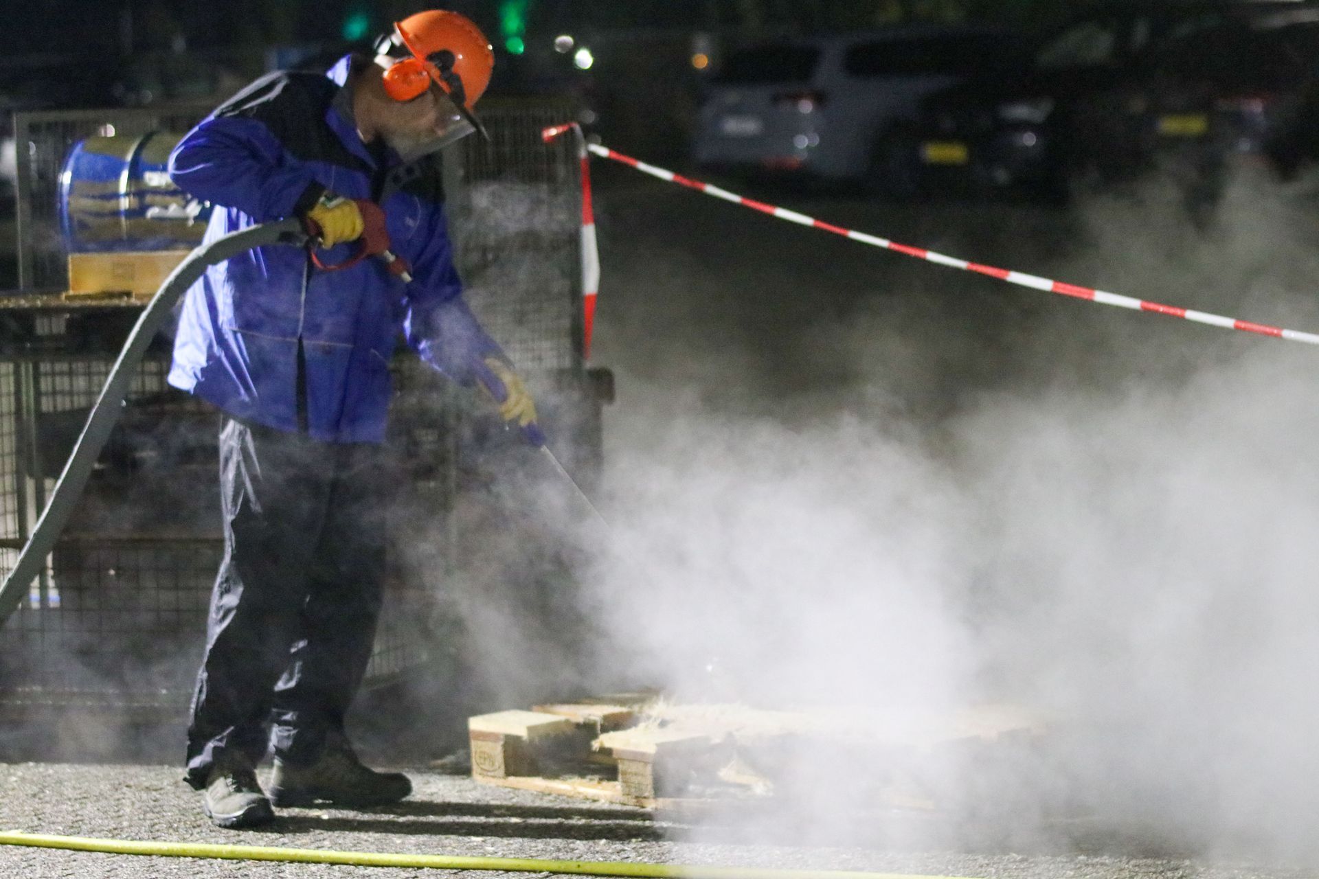 Worker in blue rain gear and orange helmet sprays water at a smoky street scene behind caution tape.