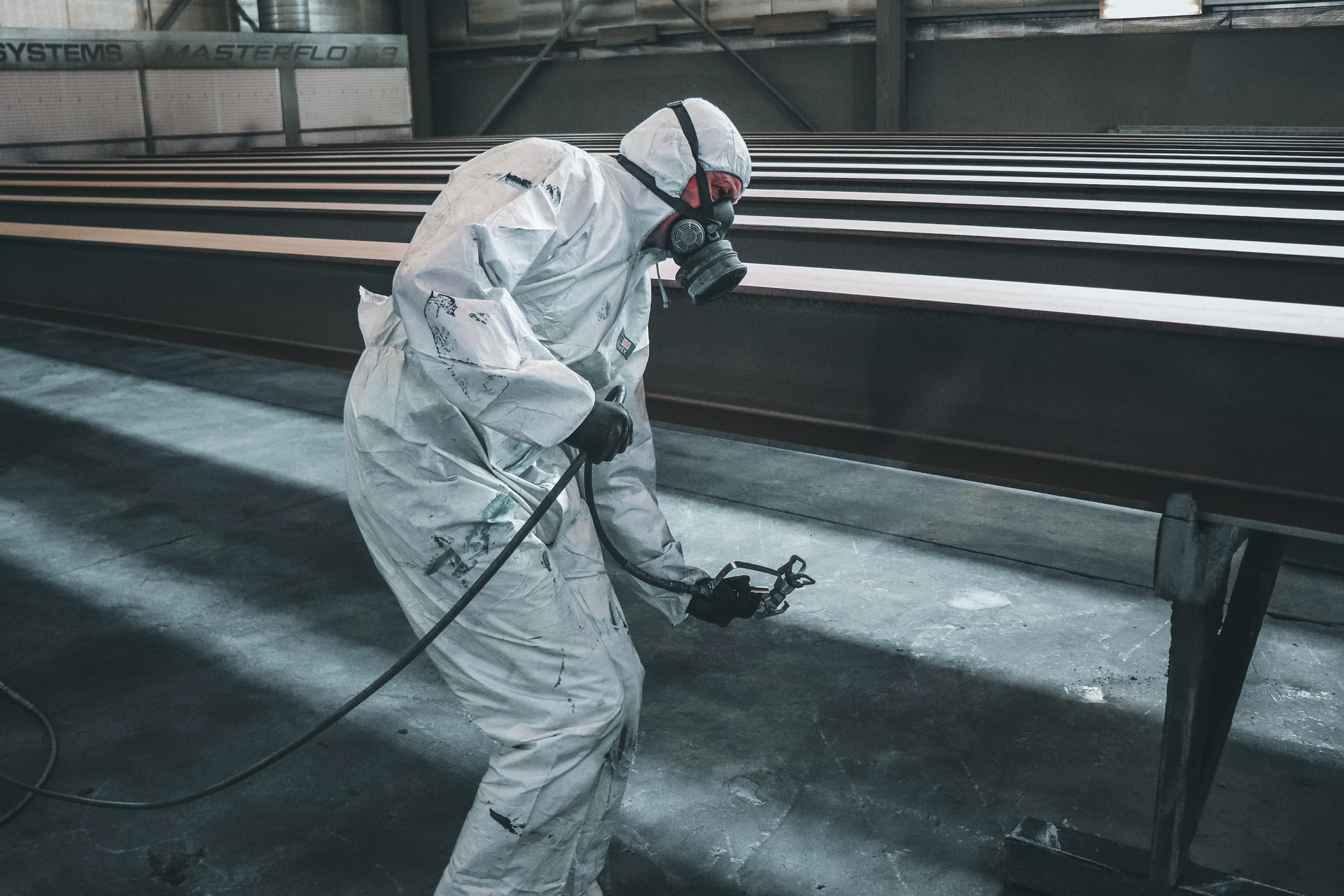 Worker in white protective suit spray-painting metal beams in an industrial facility.