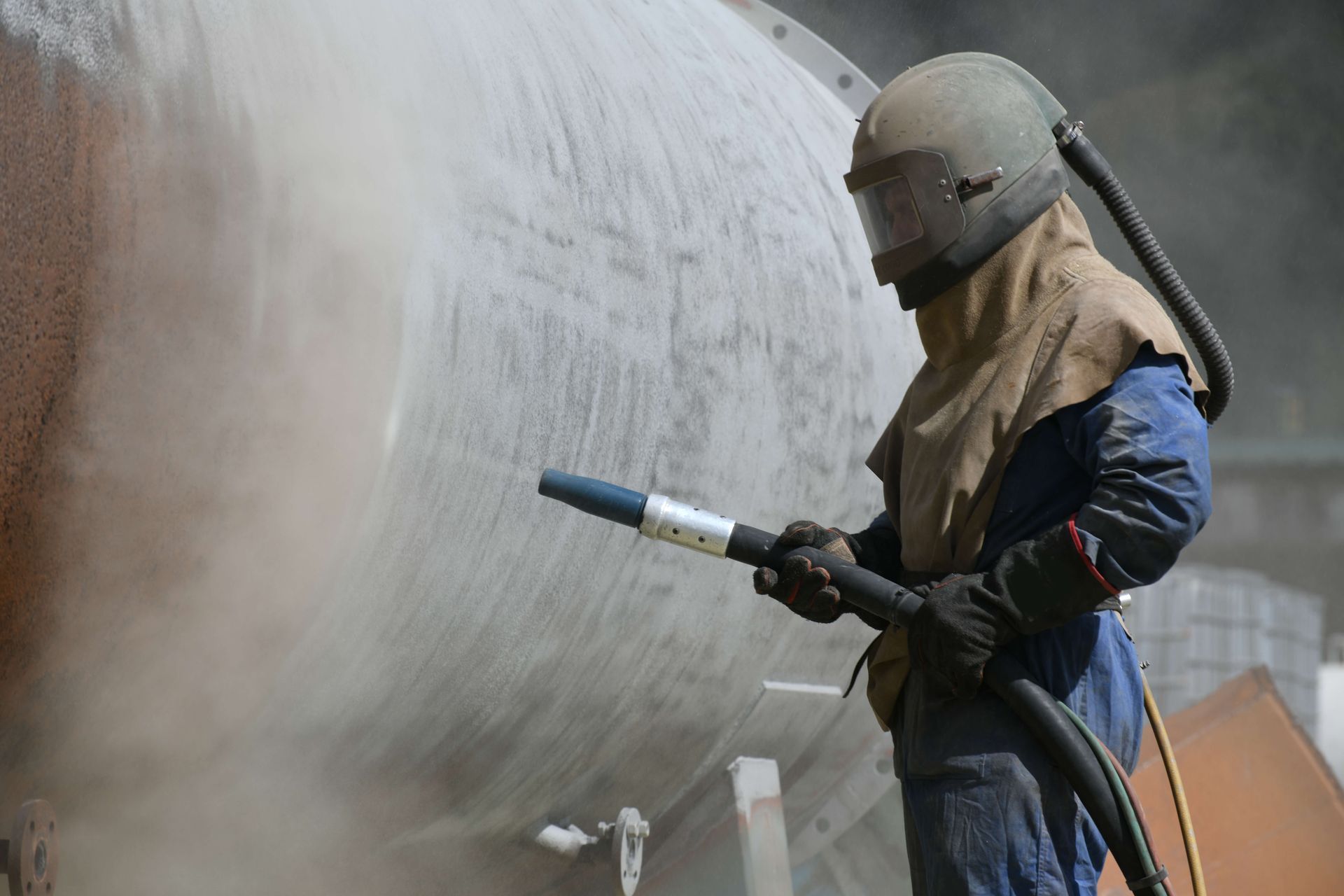 Worker in protective gear spraying a large metal tank with a high-pressure hose