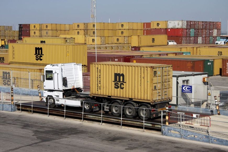 A white semi-truck carrying a yellow MSC shipping container drives over a weighbridge in a large container terminal.