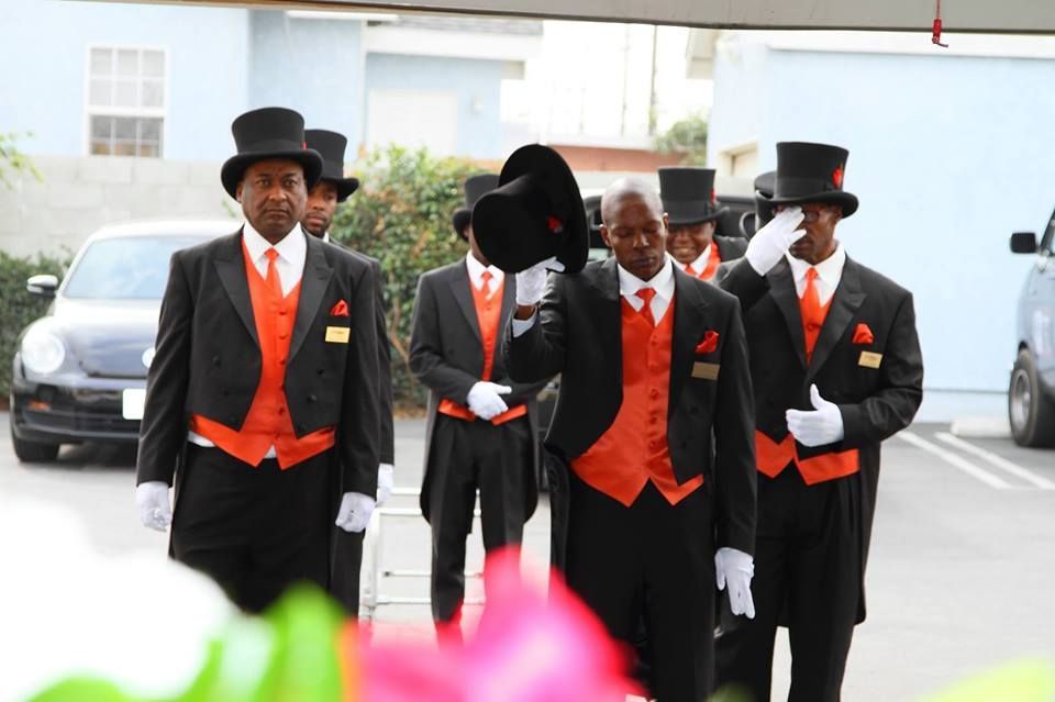 Four men in black suits with orange vests and top hats walking outdoors toward the camera