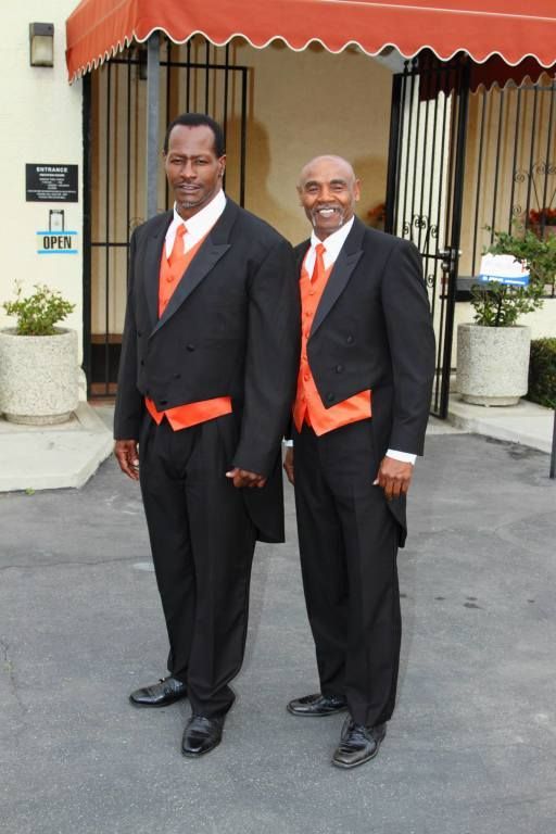 Two men in black suits with orange ties standing outside a building entrance