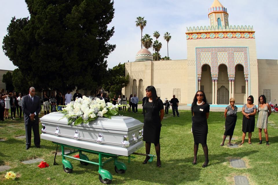White casket with flowers at an outdoor funeral service beside a mosque-like building