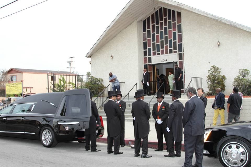 Men in dark suits gather outside a white church beside black hearses and a coffin at the entrance