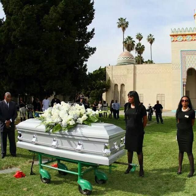 White casket on green wheeled bier at outdoor funeral, with mourners standing nearby.