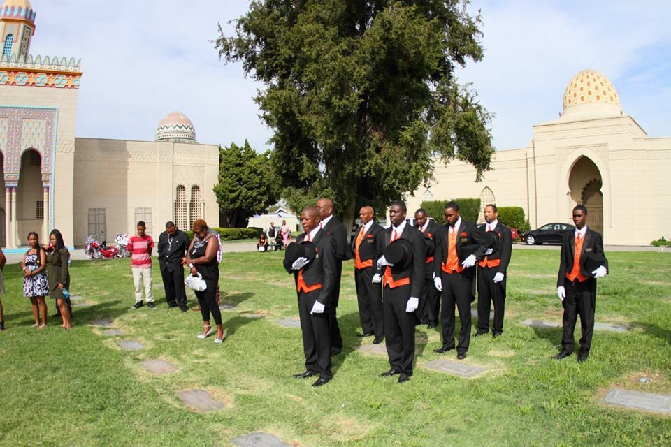 Men in black suits with orange sashes walking on grass in a courtyard near a building, with onlookers nearby