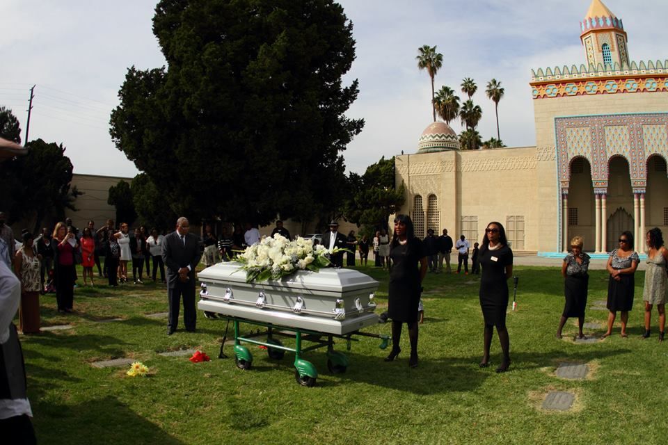 Silver casket on a lawn with mourners standing around it beside a mosque-like building