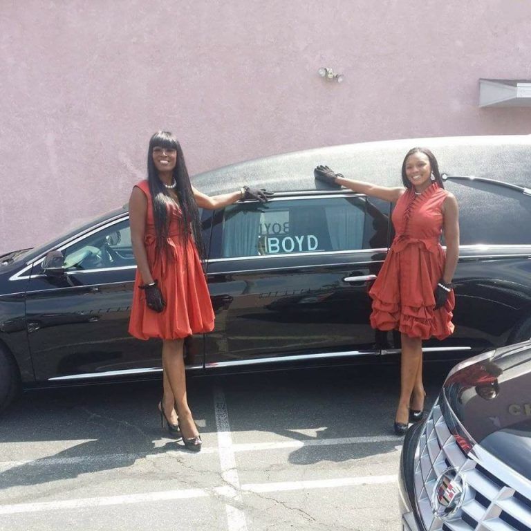 Two women in red dresses stand beside a black limousine with “BOYD” on the window.