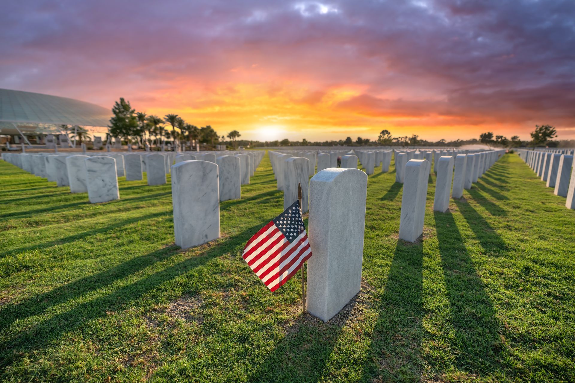 Rows of white gravestones at sunset with an American flag in a cemetery.