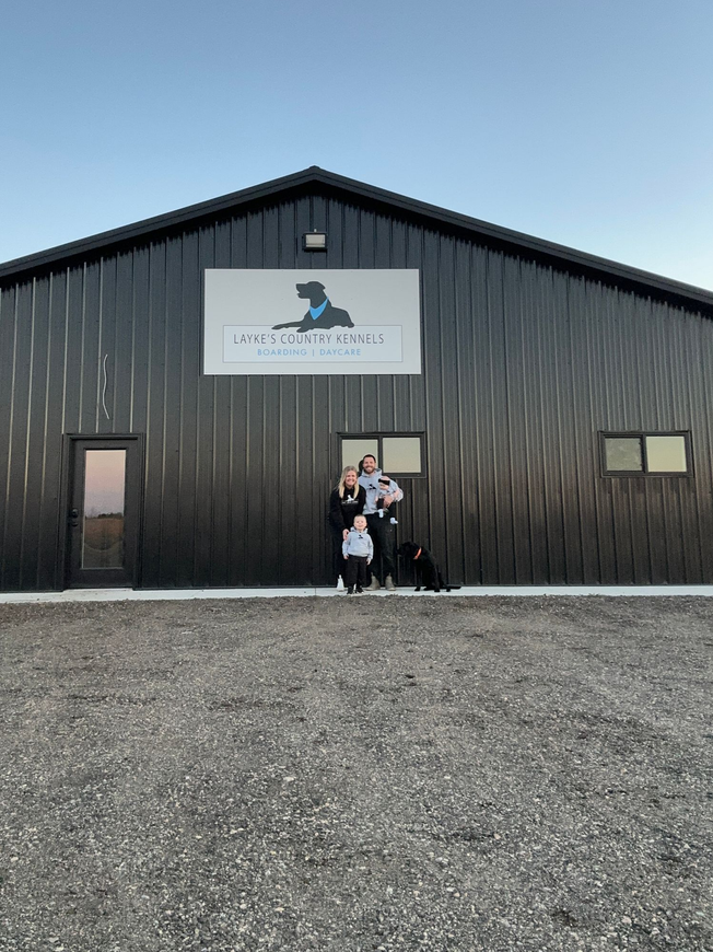 Two people and a dog stand in front of a black building with a sign. Gravel ground, blue sky.