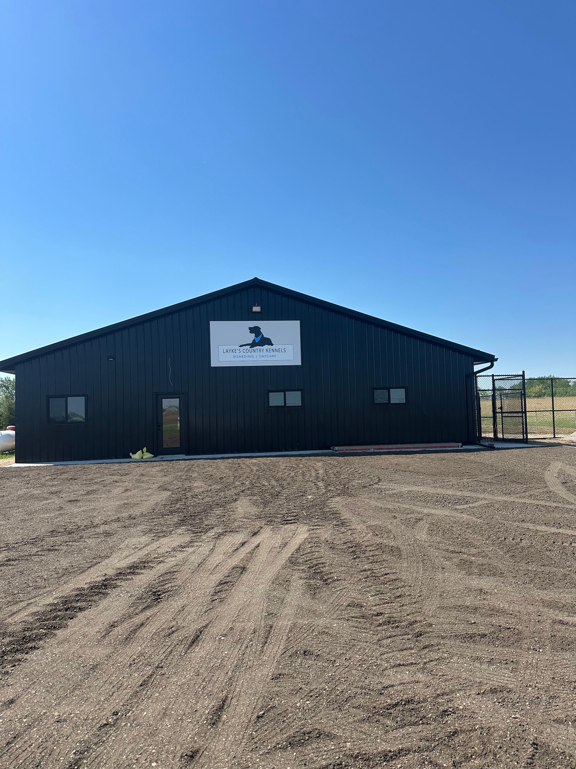 Black barn with white sign, set in a dirt lot under a clear blue sky.