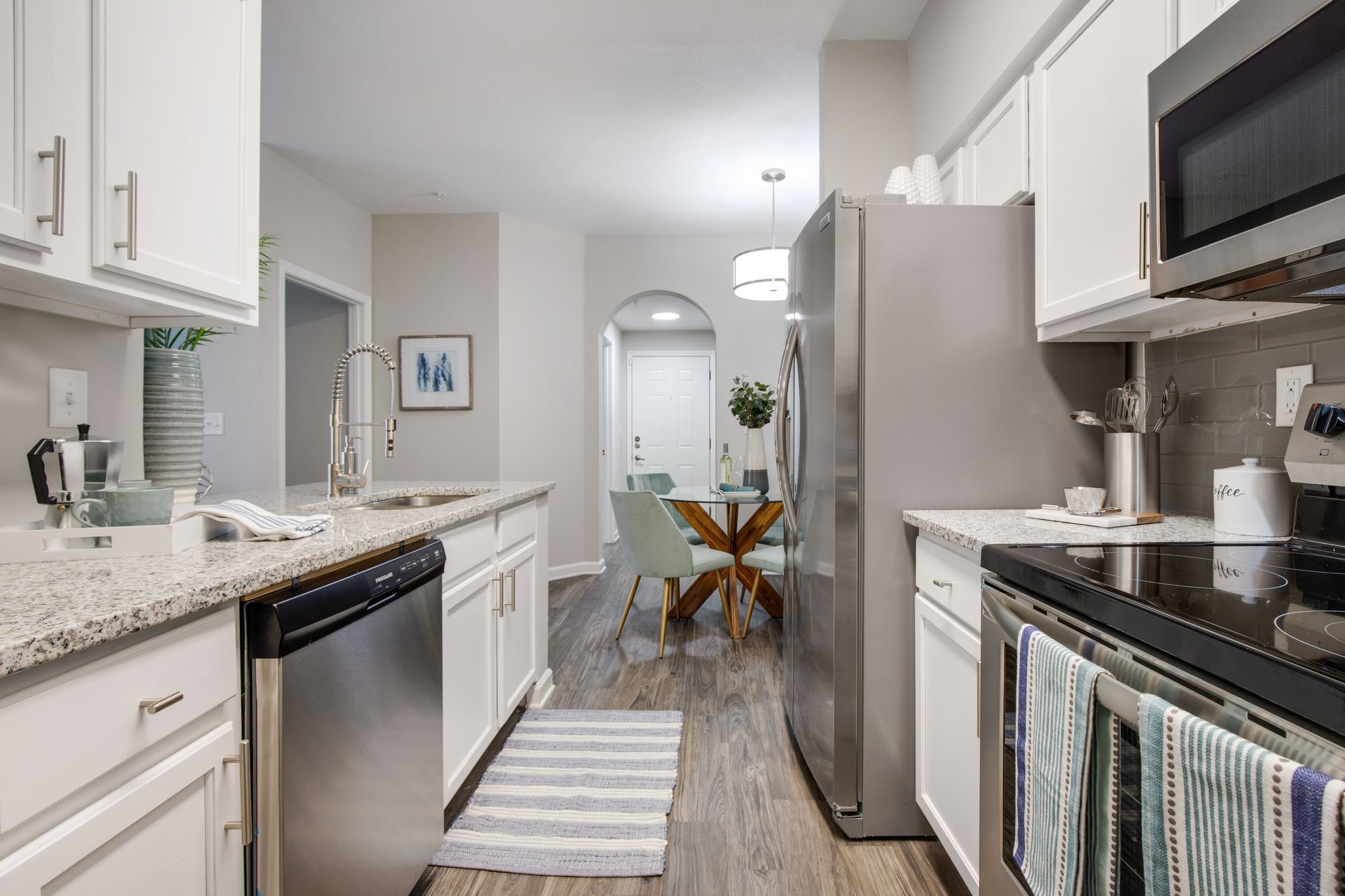 Bright galley kitchen with white cabinets, stainless steel appliances, and a small dining table at the end of The Carlyle at Godley Station apartments in Pooler, GA.