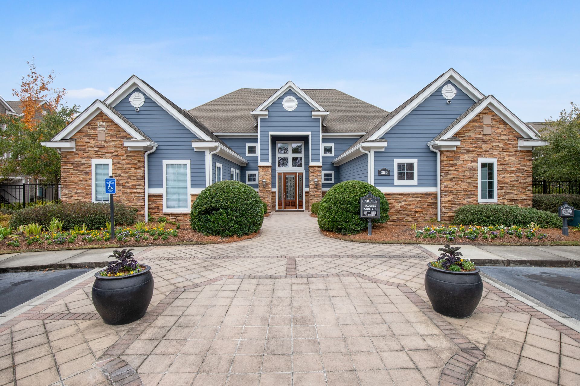 Stone-front suburban house with blue accents, a paved driveway, and two potted plants by the entrance at The Carlyle at Godley Station apartments in Pooler, GA.