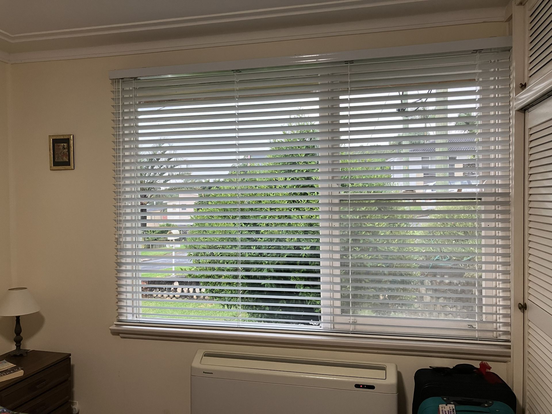 Living room with window blinds, view of greenery outside, and an air conditioner below the window