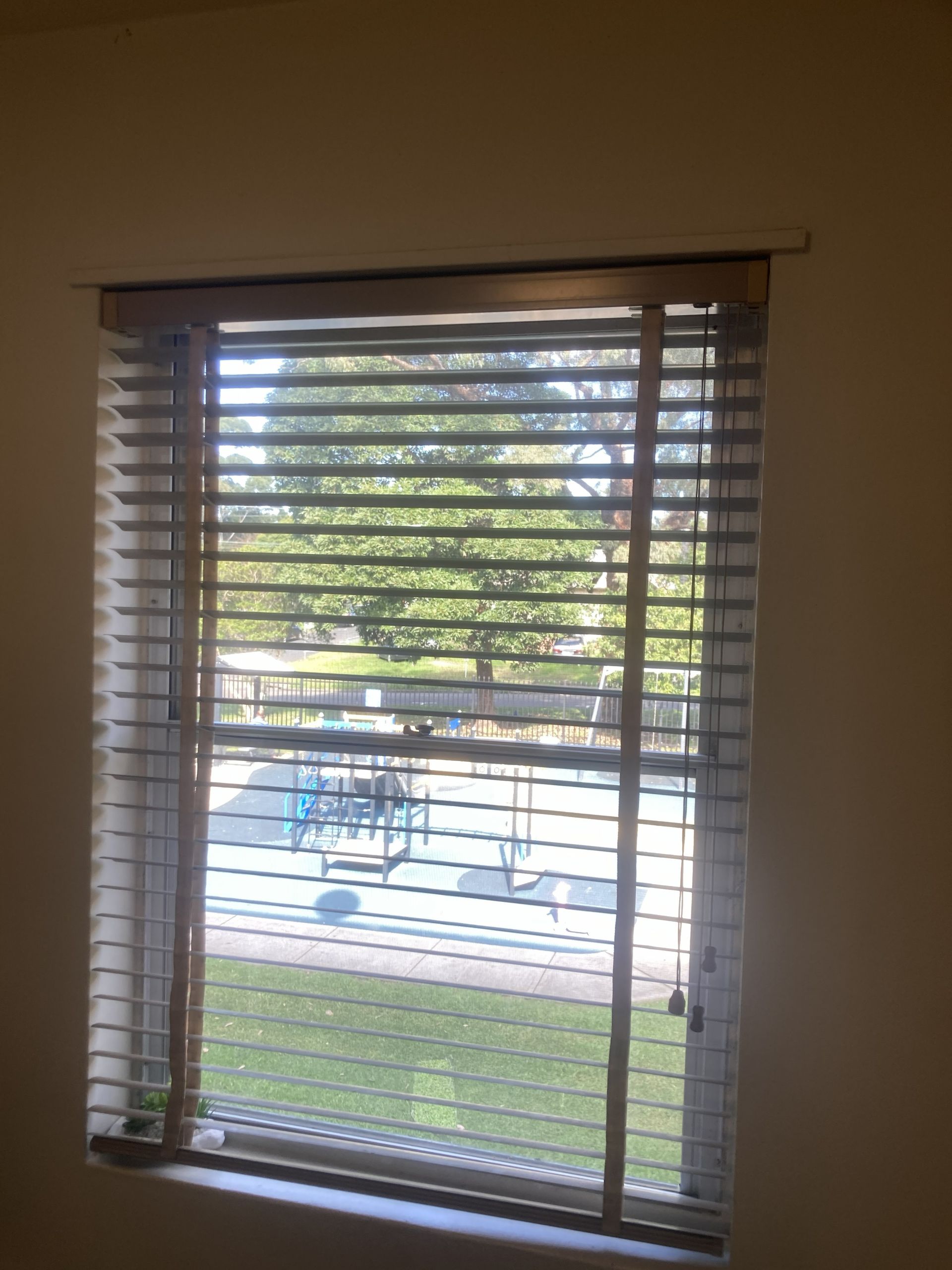 View through a window with blinds toward a patio and pool outside, with trees in the background