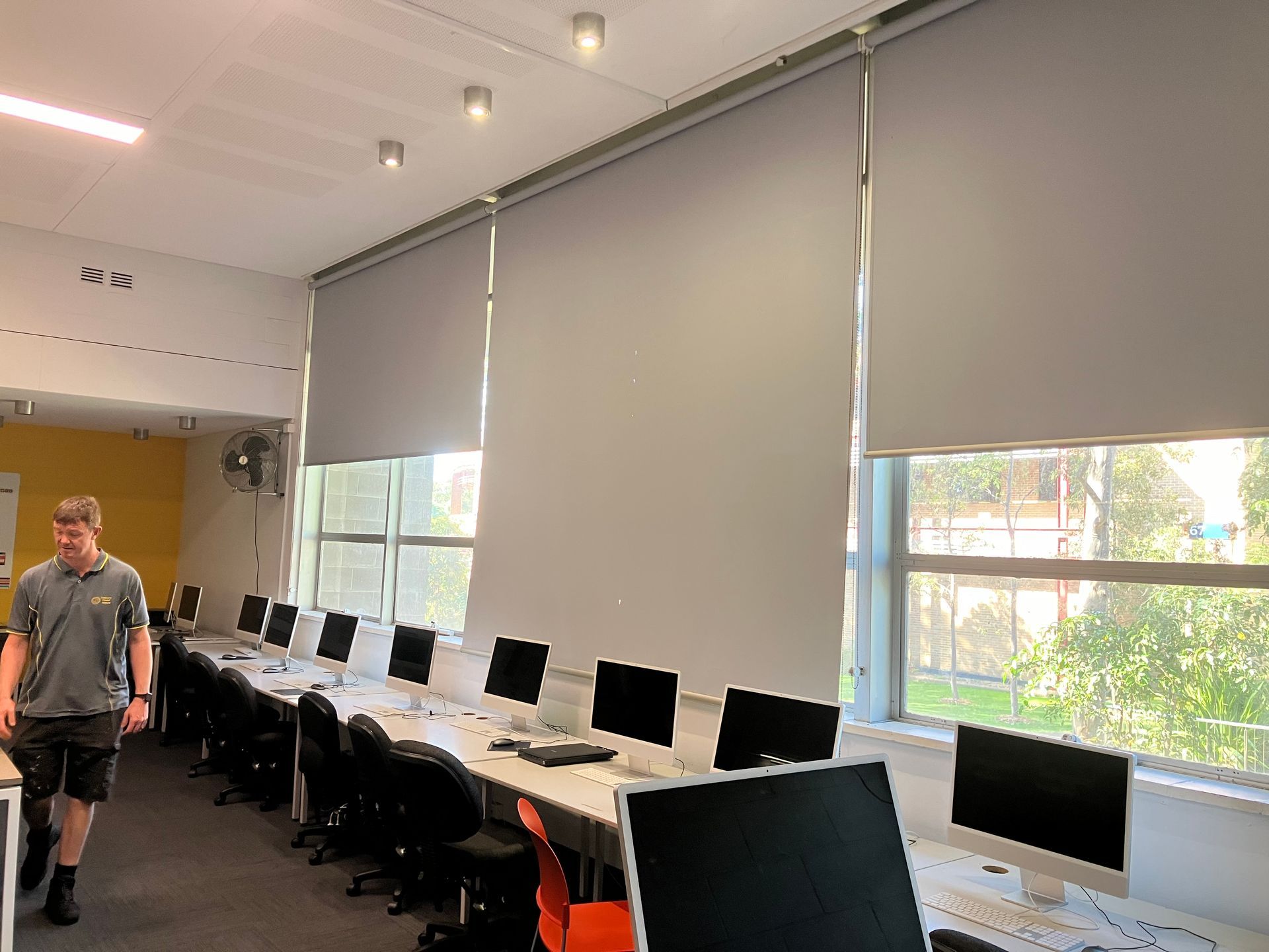 Computer lab with rows of desktop monitors and chairs by large windows; one person walks on the left.