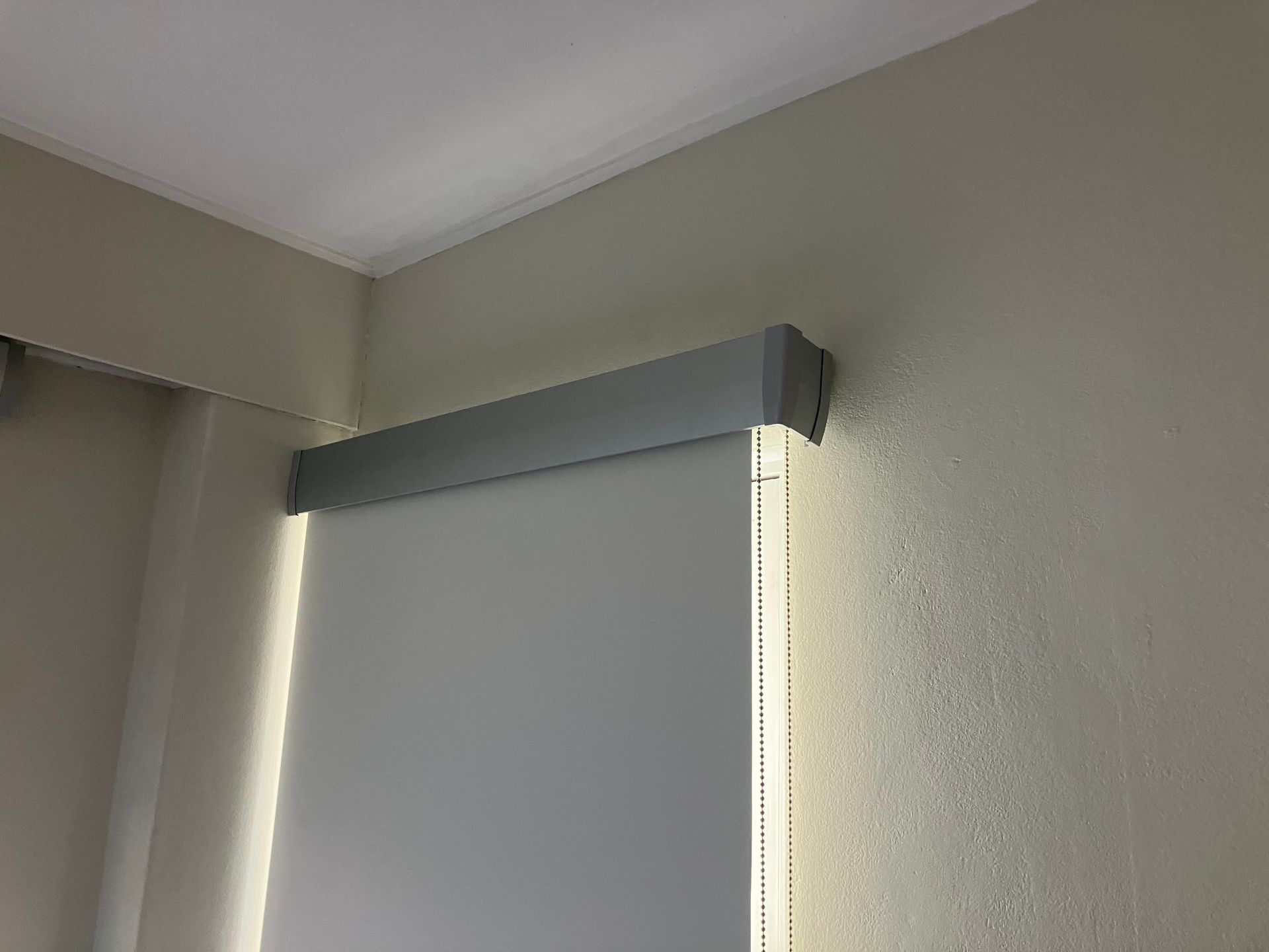 Corner of a room with a gray window blind mounted near the ceiling and a beige wall