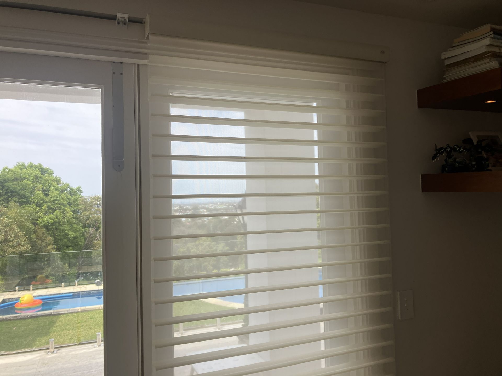 Window with white blinds overlooking a sunny backyard and pool