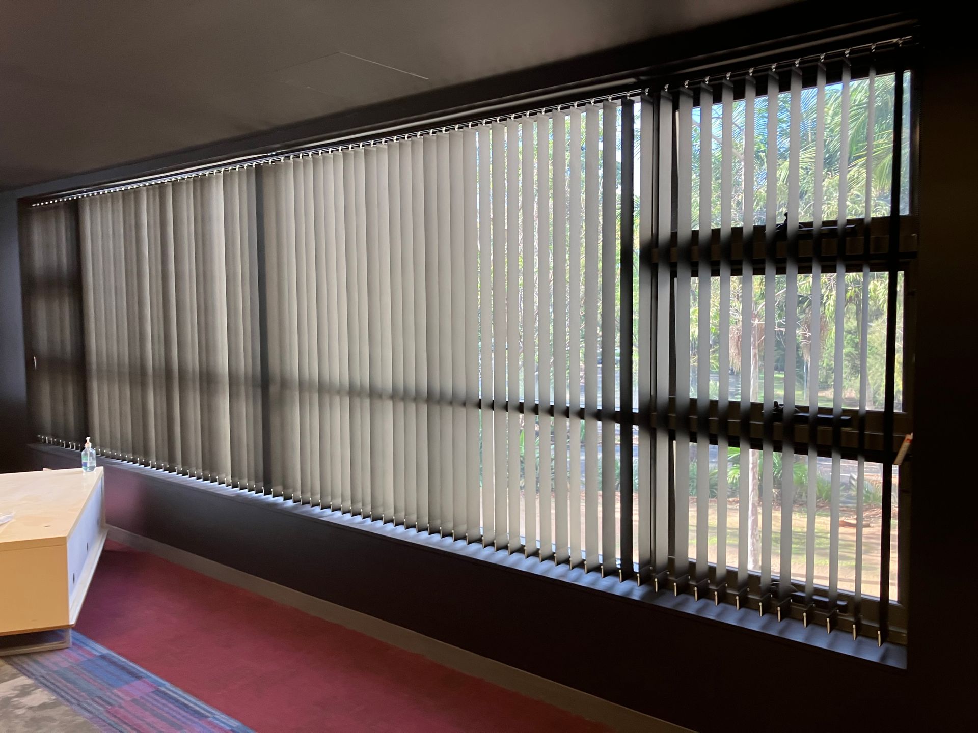 Empty room with vertical blinds covering large windows and sunlight filtering in.
