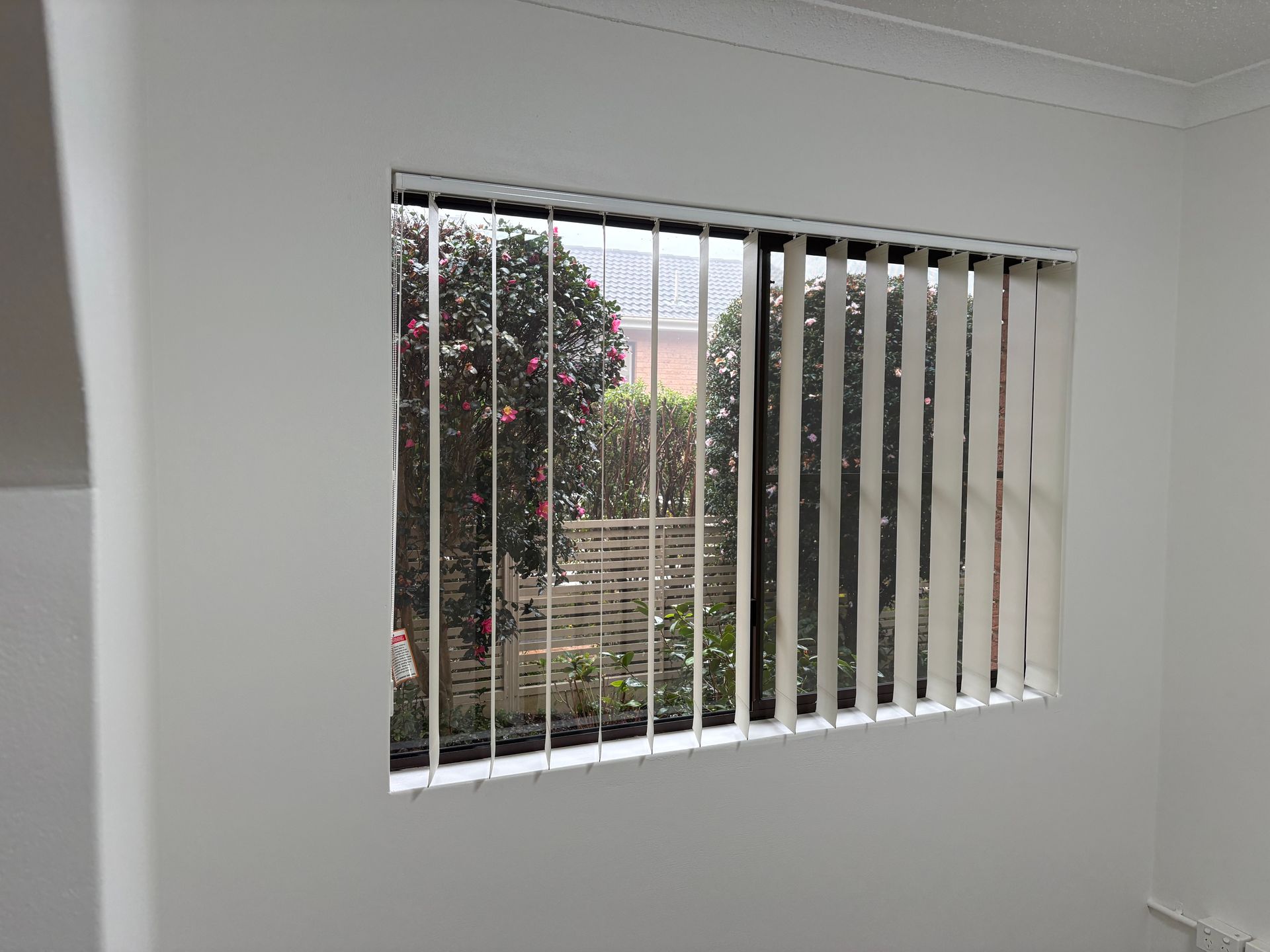 White room with a window and vertical blinds partially open, showing a garden outside