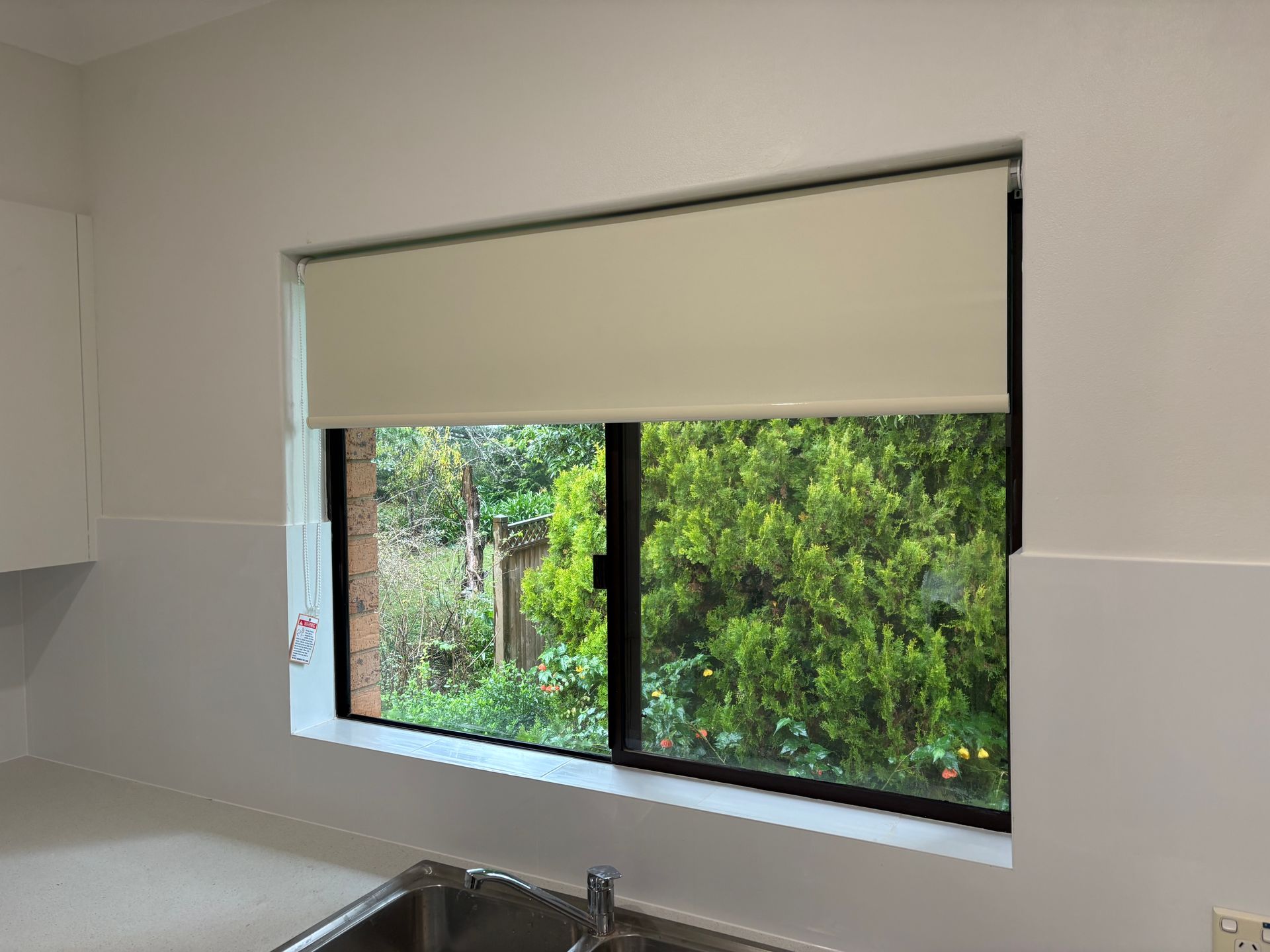 Kitchen window with white roller blind and view of green trees outside