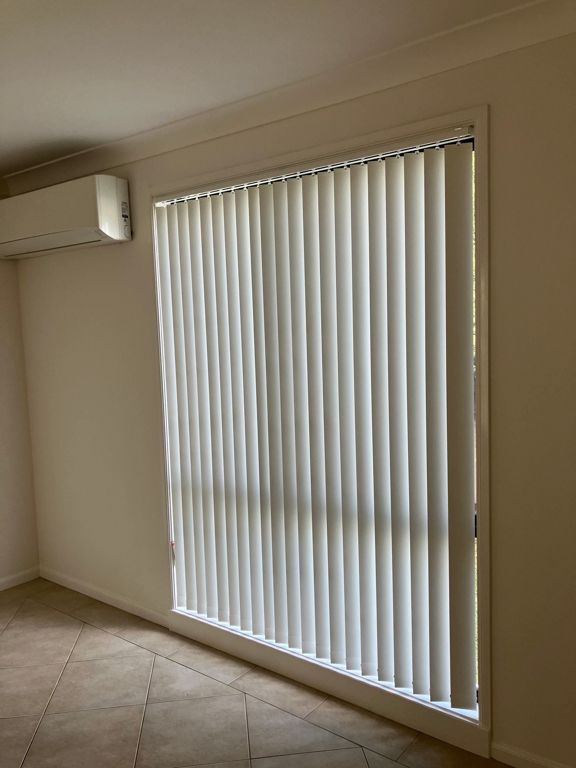 Empty room with beige walls, tiled floor, wall-mounted AC, and a large window with vertical blinds.