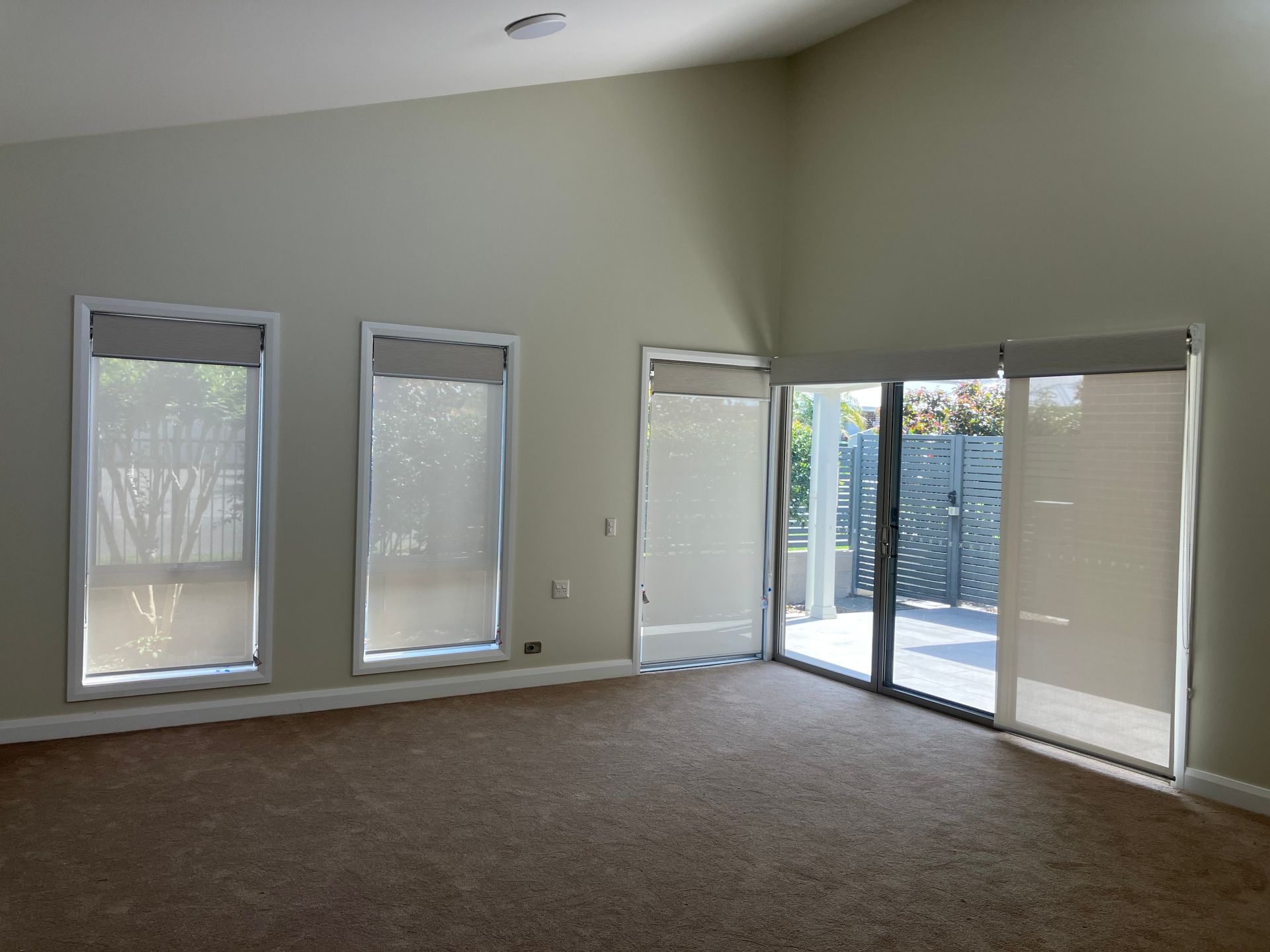 Empty living room with beige carpet, high ceiling, and tall windows and glass doors letting in daylight
