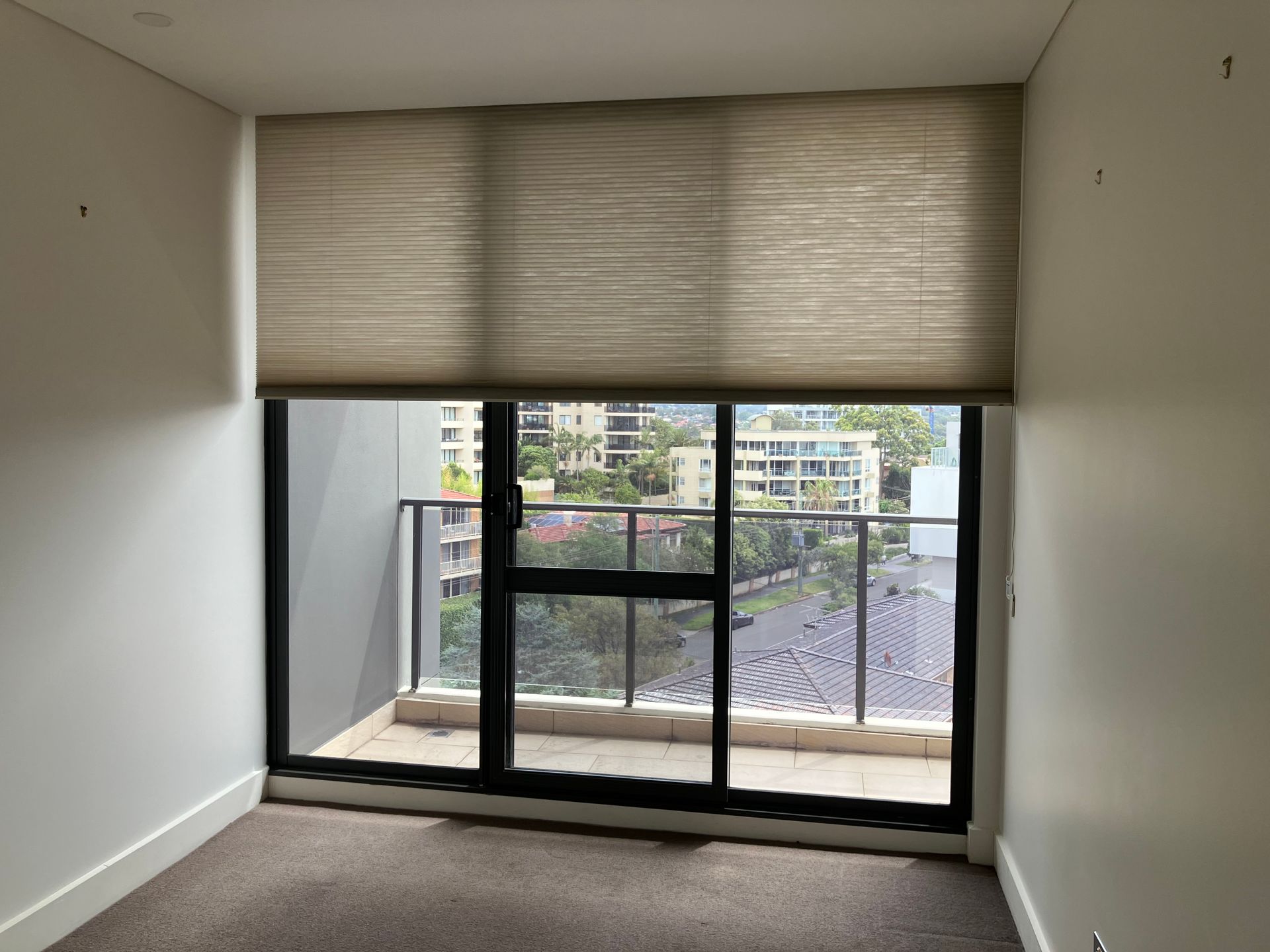 Empty room with large sliding glass doors and beige roller blind overlooking a balcony and buildings.