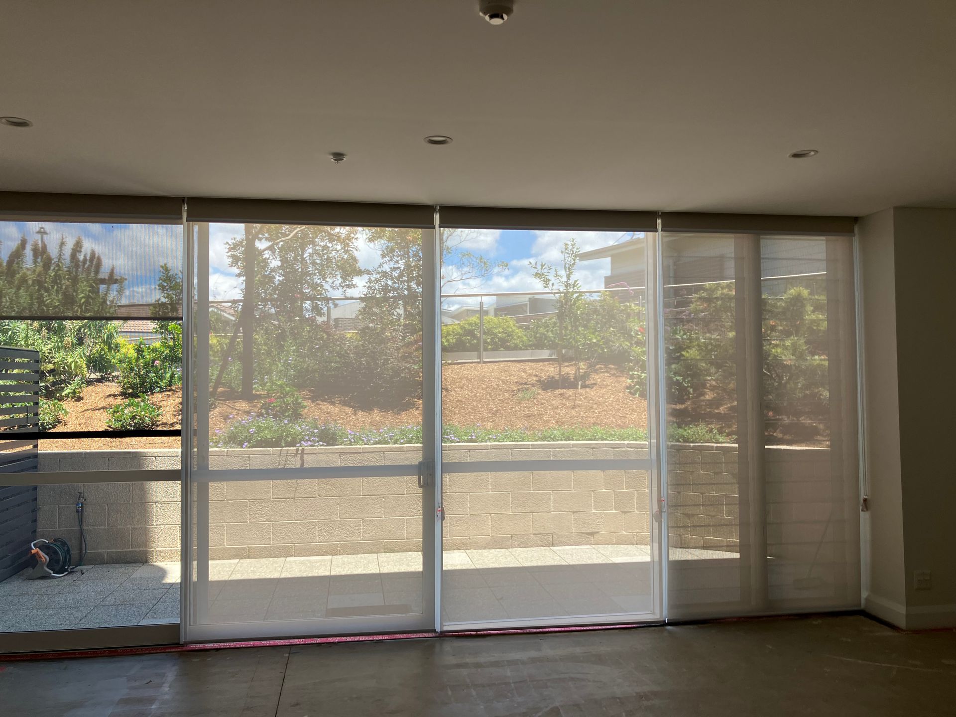 Empty room with large sliding glass doors and sheer curtains overlooking a sunny outdoor patio and trees