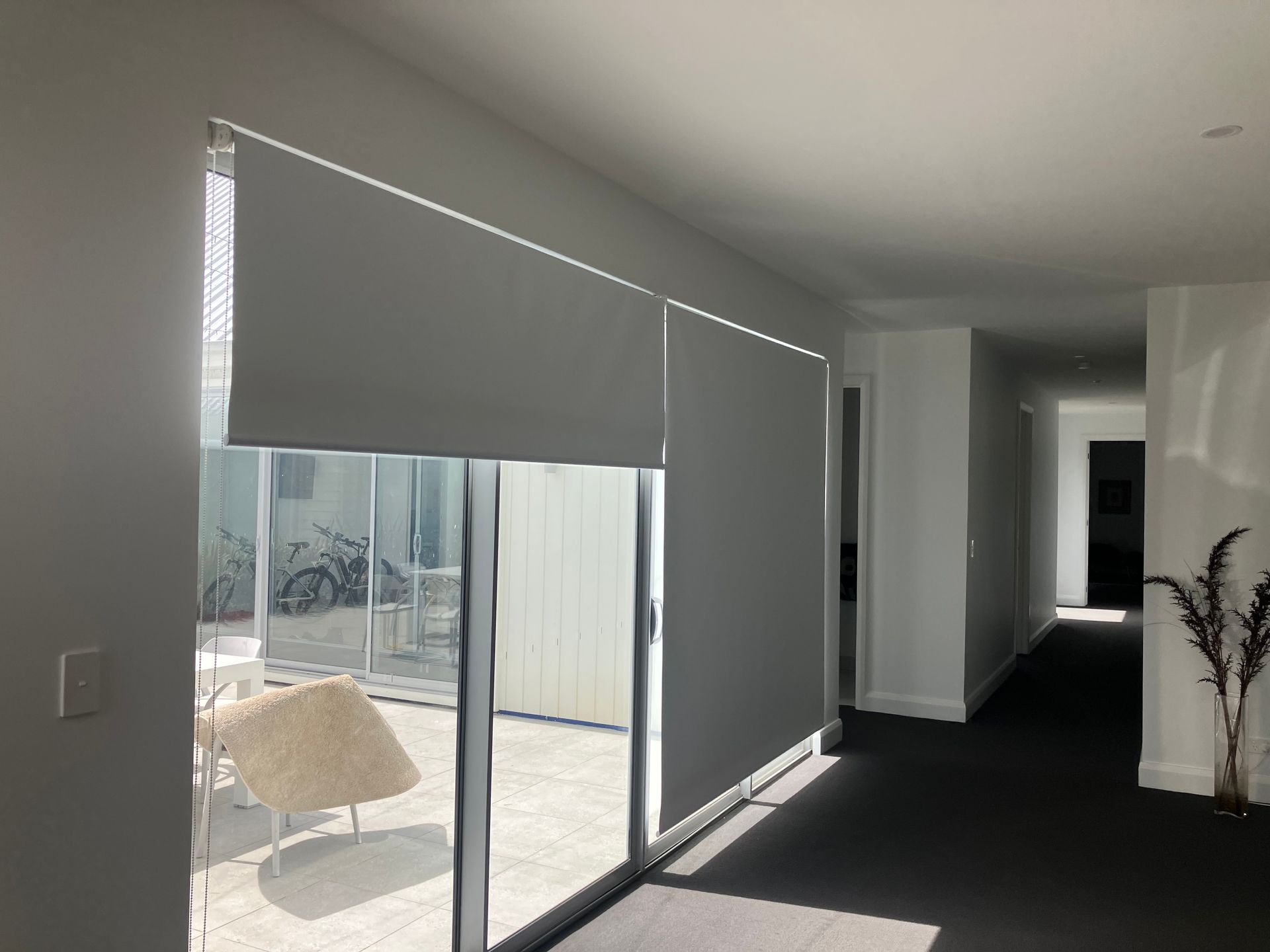Bright living room with gray blinds, sliding glass doors, and dark carpet leading to a hallway