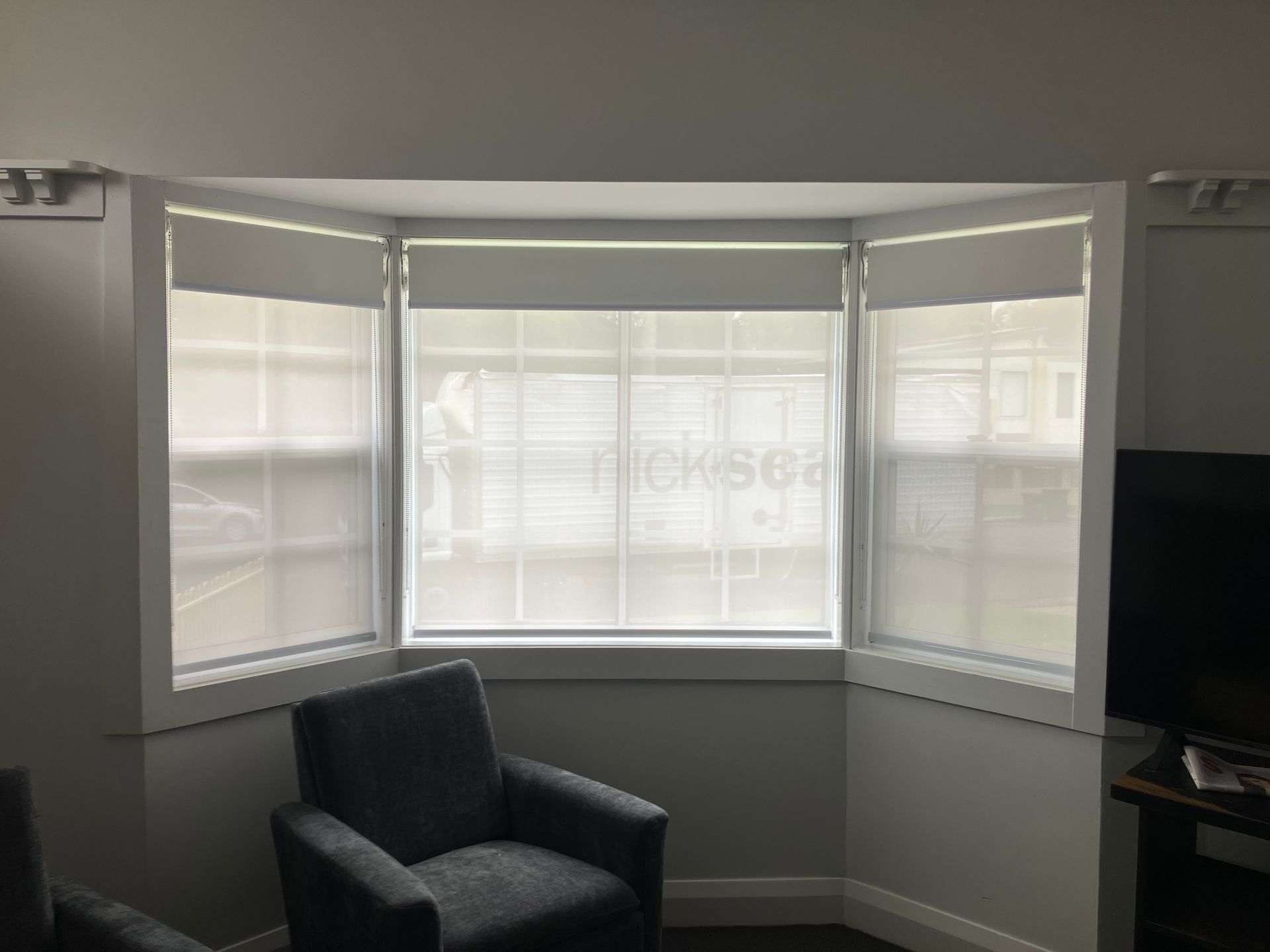 Bright bay window with white blinds, dark armchair, and a TV in a living room corner.