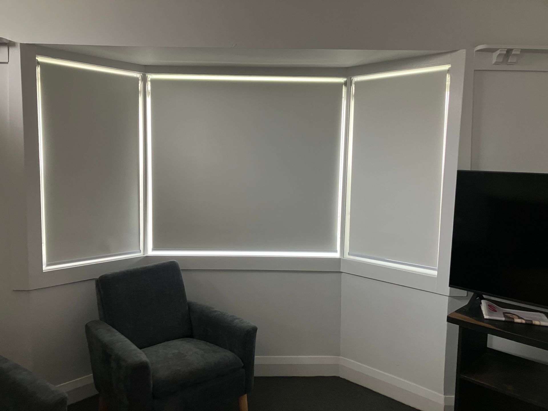 Gray corner room with three windows, a black armchair, and a TV stand on the right.