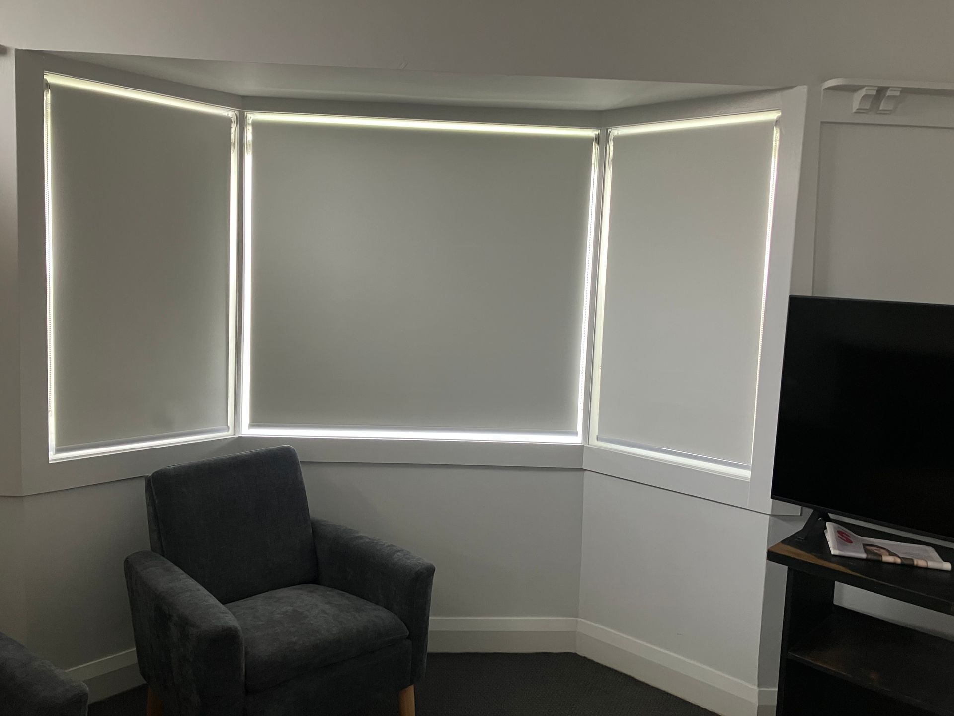 Small corner room with white blinds, a dark armchair, and a TV on a stand